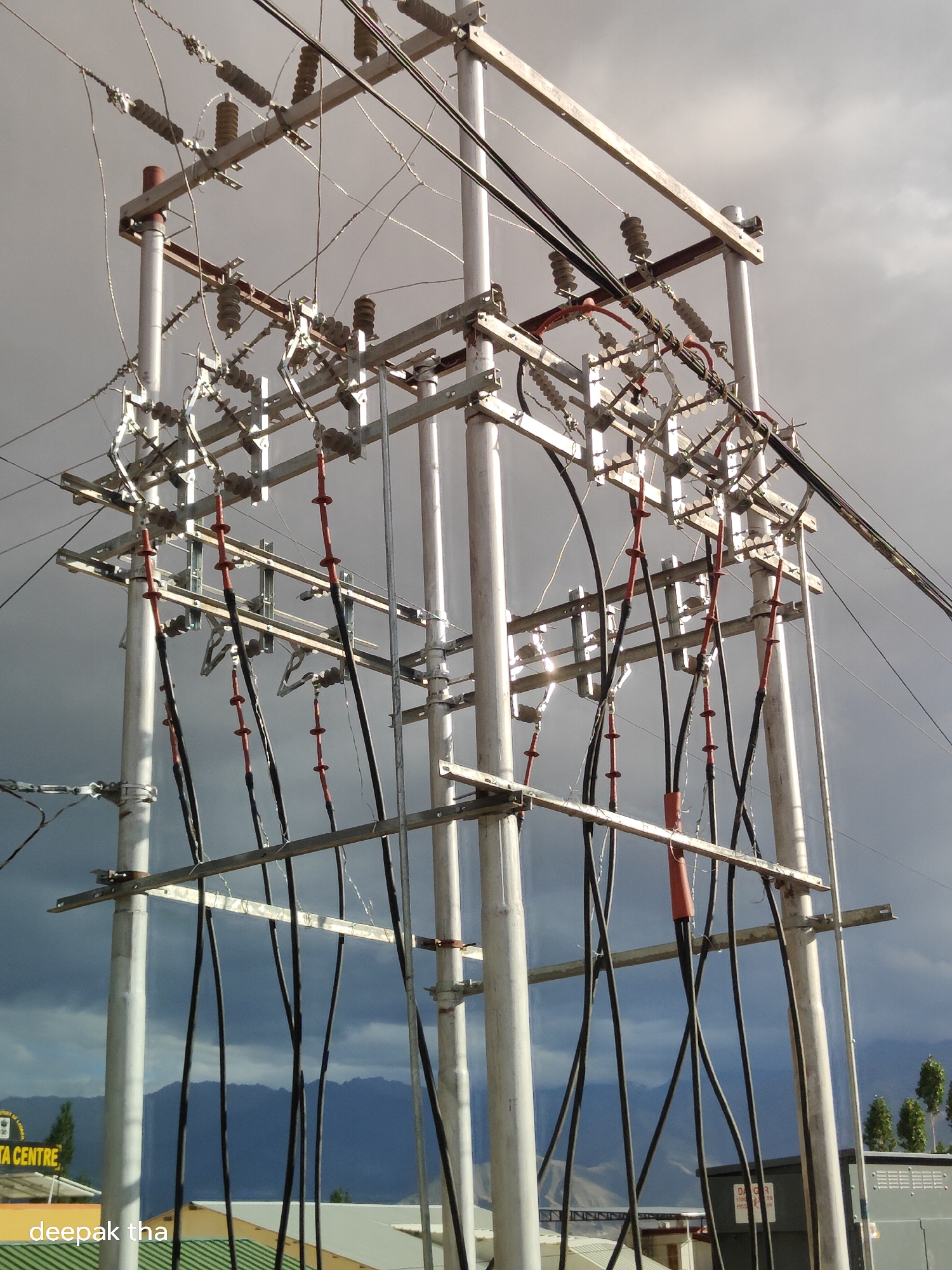 A power substation is pictured in Leh, India, under a dramatic, cloudy sky. The substation features a complex network of metal poles, crossbars, and numerous electrical wires and cables. Ceramic insulators are visible, suspending the wires. Several thick, black cables with red sections are hanging vertically from the substation's structure. In the background, mountains are dimly visible through the haze, and the roofs of buildings, including one with a sign that partially reads "DATA CENTRE," are present in the foreground. A watermark reading "deepak tha" is in the lower left corner of the image.