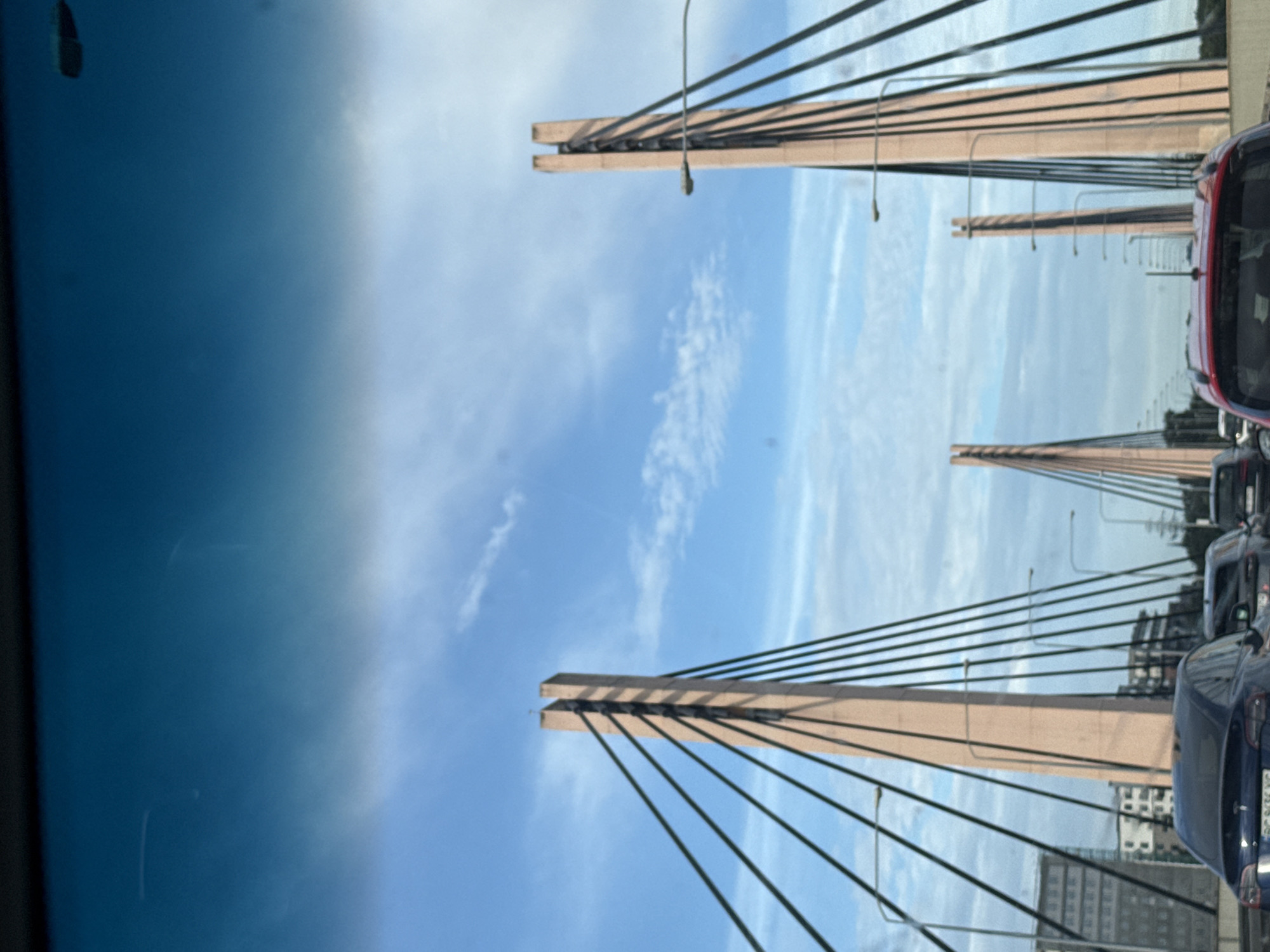 The image captures a view from a vehicle on a bridge, likely the Most Grunwaldzki (Grunwaldzki Bridge) in Wrocław, Poland. The bridge features distinctive tall, pink concrete pylons with multiple cables supporting the roadway. The sky above is blue with scattered white clouds, and there is moderate traffic on the bridge, with several cars visible in front. In the background, buildings and other infrastructure elements of the city can be seen. The perspective suggests the photograph was taken during daylight hours, possibly in the morning or late afternoon due to the angle of the light. The overall scene is one of urban transportation infrastructure under a partially cloudy sky.