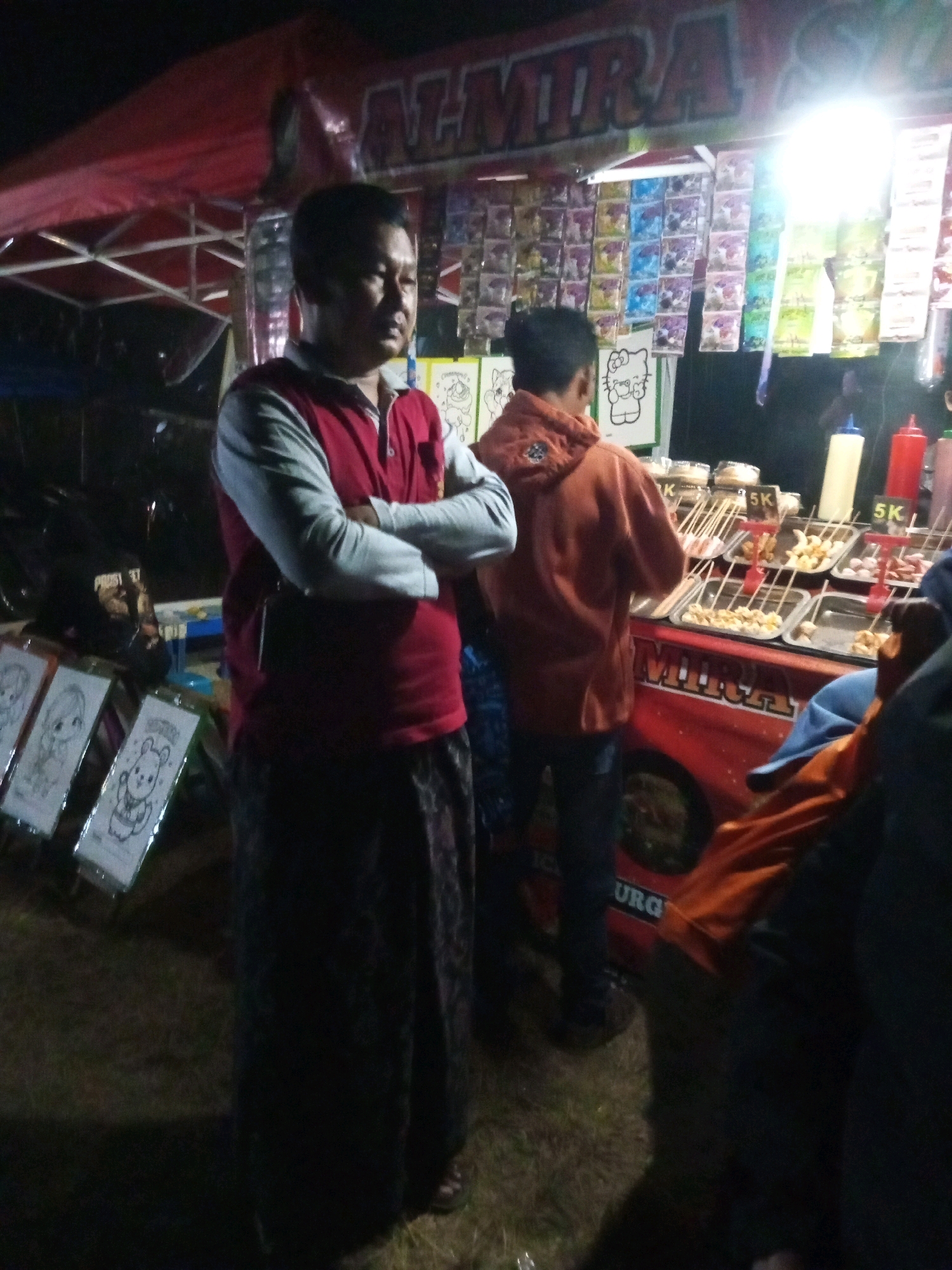 A medium shot captures an outdoor stall at night, possibly at a market or event in Ngadikerso, Indonesia. A man, appearing to be in his late 30s or early 40s, stands in the foreground, facing slightly to the right. He is wearing a red long-sleeved shirt with grey sleeves and a dark, patterned sarong. His arms are crossed over his chest. To his right, another person, likely a young man, faces away from the camera, wearing an orange hooded sweatshirt and dark pants. This person is looking towards the stall.
The stall itself is set up under a red tent with a metal frame. Behind the second person, shelves are lined with various items, including packets of what appear to be snacks or instant noodles, displayed in rows. The stall counter displays trays of food on skewers, resembling satay or grilled snacks, with prices indicated by "5K" (likely 5,000 Rupiah). Two large bottles, one white and one red, are visible on the counter, possibly containing sauces or condiments. The stall's facade is decorated with a red surface featuring the word "ALMIRA" in large, white letters. To the left of the main stall area, several framed pictures or posters are displayed, including line drawings of characters like Hello Kitty and other cartoon figures. The ground appears to be covered in dry grass and dirt. The scene is illuminated by artificial light, creating a nighttime atmosphere.