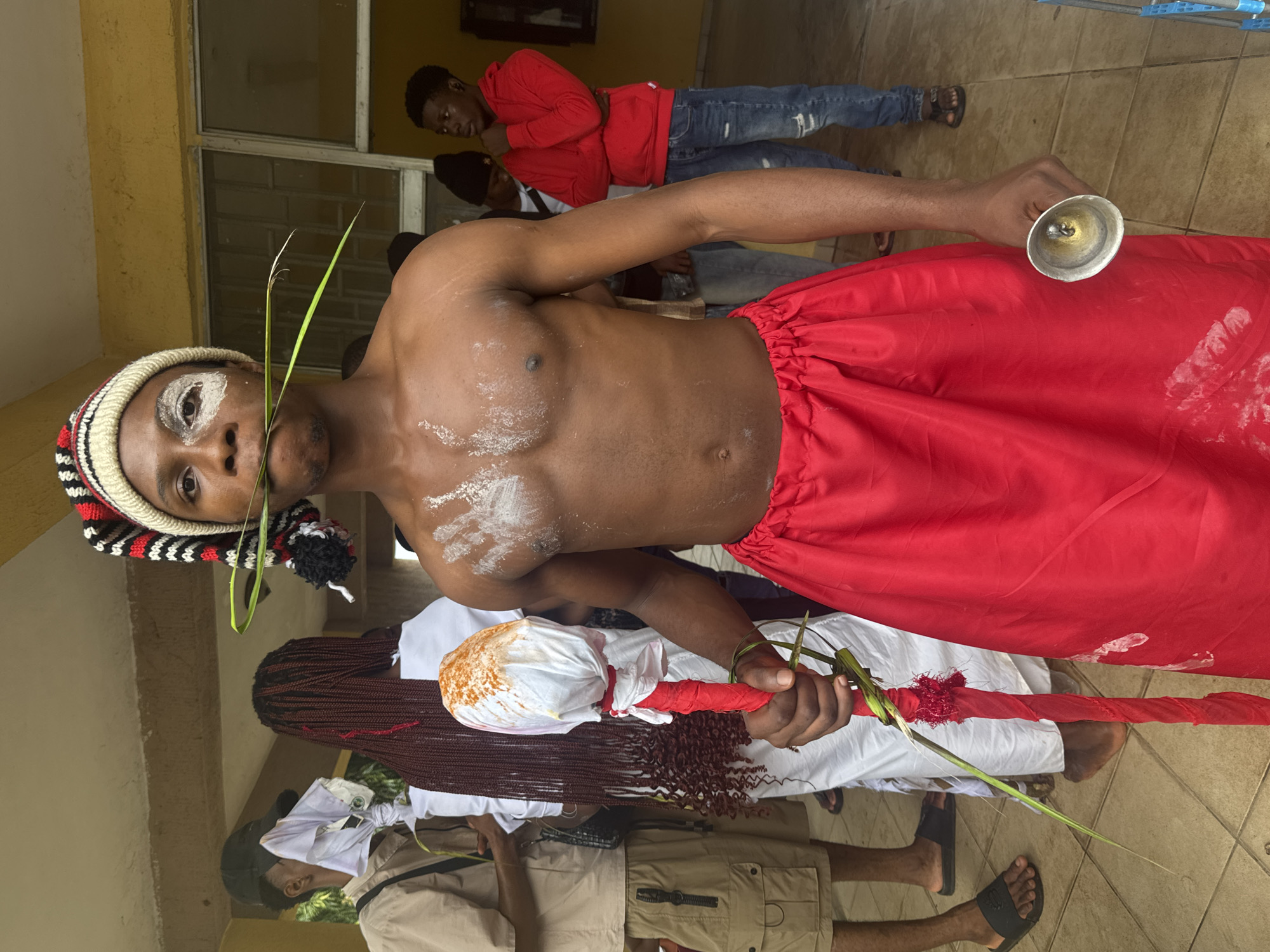 In Ihiagwa, Nigeria, a young Black male, adorned with white powder on his chest and face, holds a staff wrapped in red fabric and white cloth, with a red beaded decoration at the top. A green leaf stem is held in his mouth, and he wears a red skirt and a striped knit cap. In the background, other individuals are visible, including a person in a white garment with long, dark braids, and another in a red hoodie and jeans. The scene appears to be outdoors, near a building with a covered walkway.