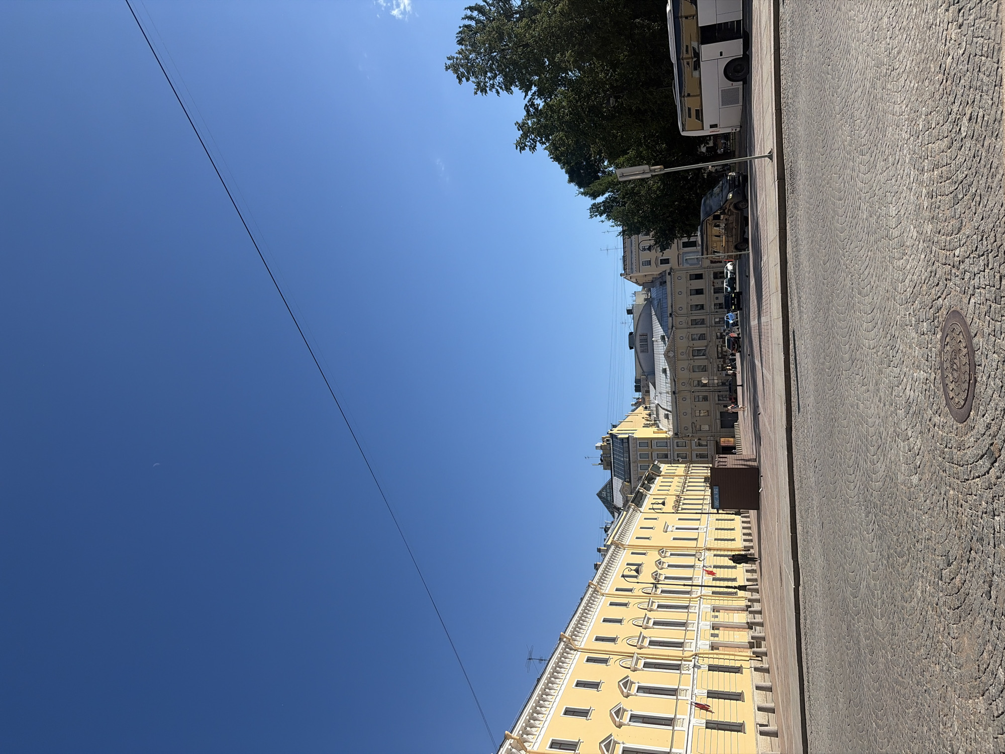 This image captures a sunlit street scene in Saint Petersburg, Russia, likely on a clear day. The foreground is dominated by a cobblestone street with a circular manhole cover. A wide, paved area separates the street from a row of historic buildings.
The buildings lining the street are predominantly yellow with white architectural details, featuring numerous windows. The architecture suggests a classic European style. Flags hang from the facades of some buildings, indicating official or cultural significance. A few small wooden kiosks are present along the pedestrian walkway in front of the buildings.
Several vehicles are visible. A white bus with open doors is parked on the right side of the frame. A dark-colored van is situated further down the street. Other cars are also present, suggesting active city life.
A few individuals are walking on the pedestrian area, appearing as small figures in the distance. One person is walking towards the left along the yellow building, while others are more spread out.
The sky is a clear, deep blue with a visible crescent moon in the upper left portion of the image and a thin power line stretching across it. Trees with lush green foliage are visible on the right side, partially obscuring some of the buildings. The overall impression is one of a vibrant urban environment bathed in sunlight.
