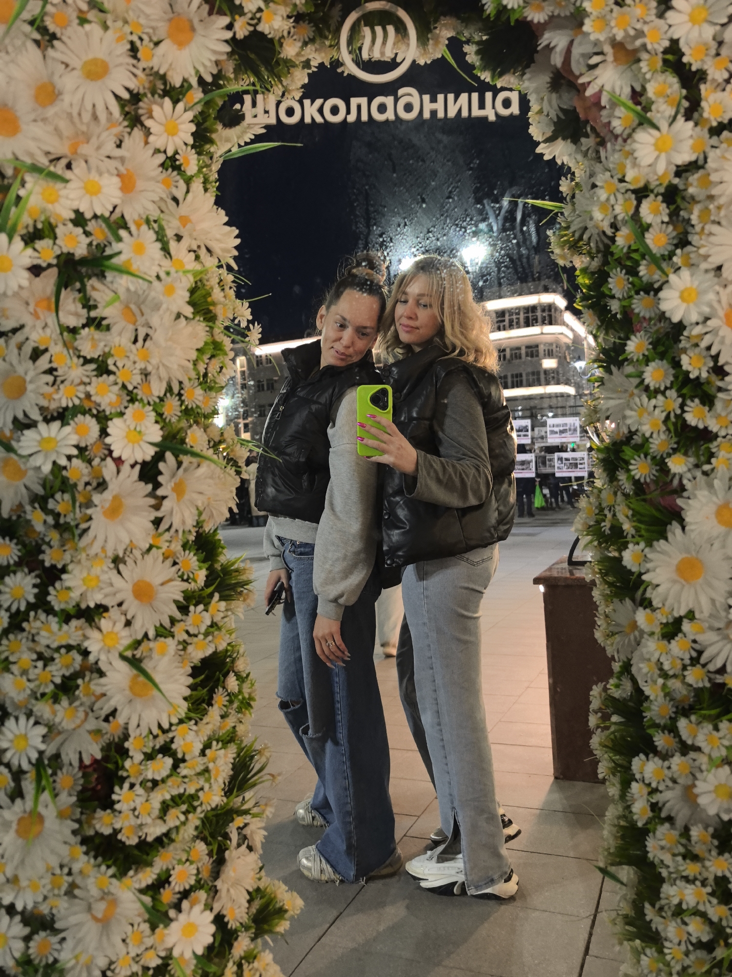 Two women are posing for a selfie in front of a mirror. The mirror is surrounded by a flower arrangement of white daisies. The location is likely Nizhny Novgorod, Russia, based on the Cyrillic text "Шоколадница" above the mirror. The woman on the left is wearing a black vest over a gray sweatshirt, blue jeans with a rip in the knee, and silver sneakers. She has her hair in a bun. The woman on the right is also wearing a black vest over a gray sweatshirt, light blue jeans with a slit on the ankle, and white sneakers. She has blonde curly hair. She is holding a green phone. The background shows a night city scene with buildings and lights. The mirror surface has water droplets.
