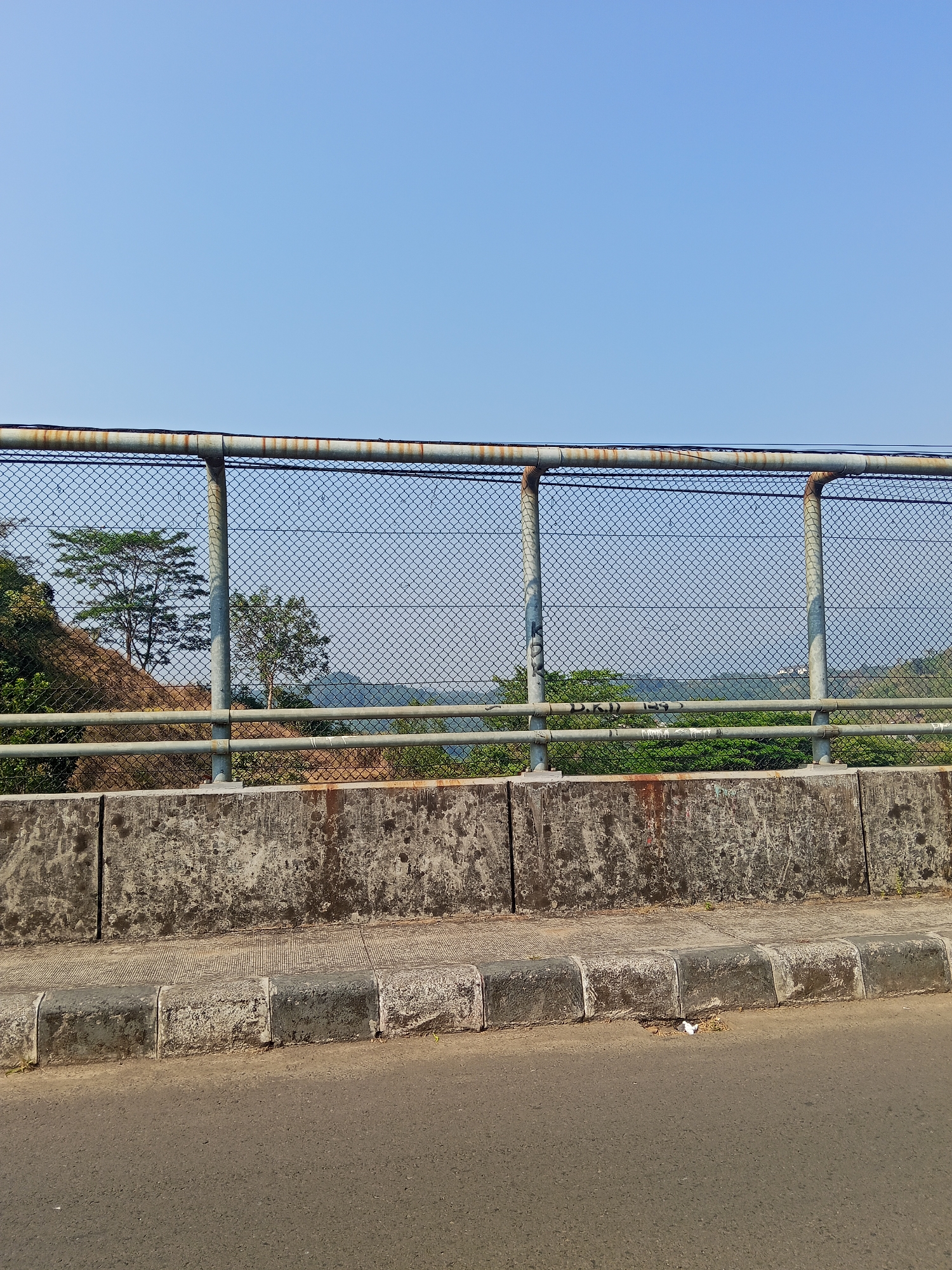The image depicts a view from what appears to be a bridge or elevated road in Semarang, Indonesia. In the foreground, a paved road runs horizontally, bordered by a concrete curb and a concrete barrier. Above the barrier, a chain-link fence runs the length of the scene, supported by metal posts. Beyond the fence, a natural landscape is visible, featuring a body of water in the distance, surrounded by rolling hills and lush green vegetation. Several trees are scattered across the terrain. The sky above is clear and blue. The overall impression is one of an urban infrastructure element overlooking a natural environment.