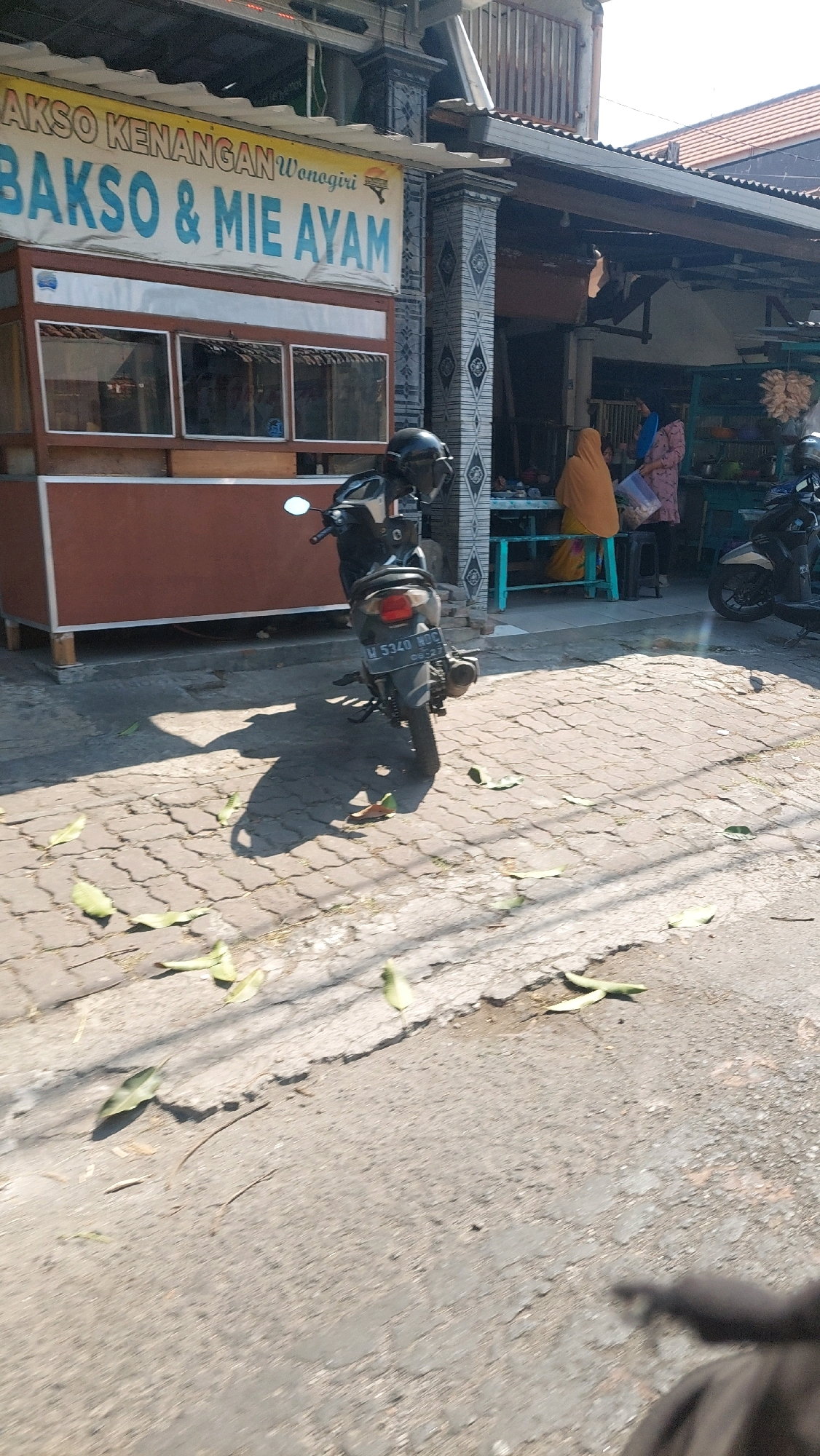 A medium shot captures a sunlit street scene in Sidoarjo, Indonesia, with a food stall named "BAKSO & MIE AYAM Wonogiri" dominating the left side. The stall features a maroon-colored counter and windows displaying its interior. A black motorcycle is parked in front of the stall, casting a long shadow on the cobblestone pavement. Several dry leaves are scattered across the ground. In the background, a few people are seen gathered around tables outside what appears to be another establishment. The overall environment suggests a typical street scene in a bustling Indonesian city.
