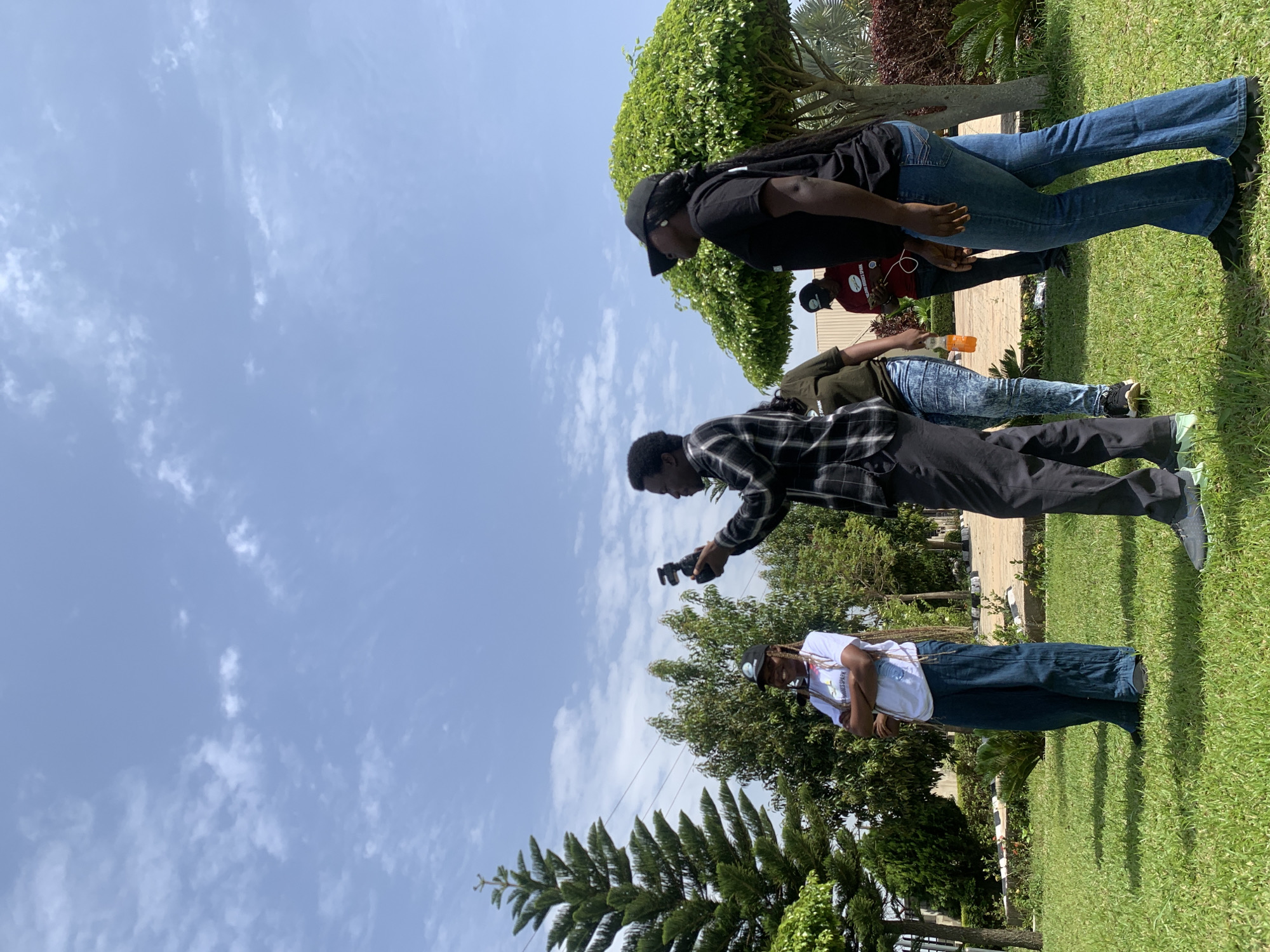 The image depicts a group of people outdoors in a grassy area, possibly a park or a garden, under a partly cloudy sky. The scene is likely set in Jos, Nigeria, given the context of previous requests.
In the foreground, a person in a black and white checkered shirt is holding a camera and appears to be taking a photo of a young woman standing to their left. The woman is wearing a white t-shirt with some text and graphics, a black cap, and wide-leg blue pants. She has her arms crossed.
To the right of the checkered-shirted person, a woman with a black cap and a black t-shirt is looking away from the camera. She is wearing blue jeans. Behind her, another woman in a green t-shirt and blue jeans is visible, holding an orange beverage. Further in the background, a person in a red shirt is seen looking at a phone.
The environment features well-maintained green grass, several trees of varying sizes and types, and some foliage. The large, conical evergreen tree on the left is a prominent feature. The sky is a light blue with scattered white clouds. The overall impression is of a casual outdoor gathering on a pleasant day.