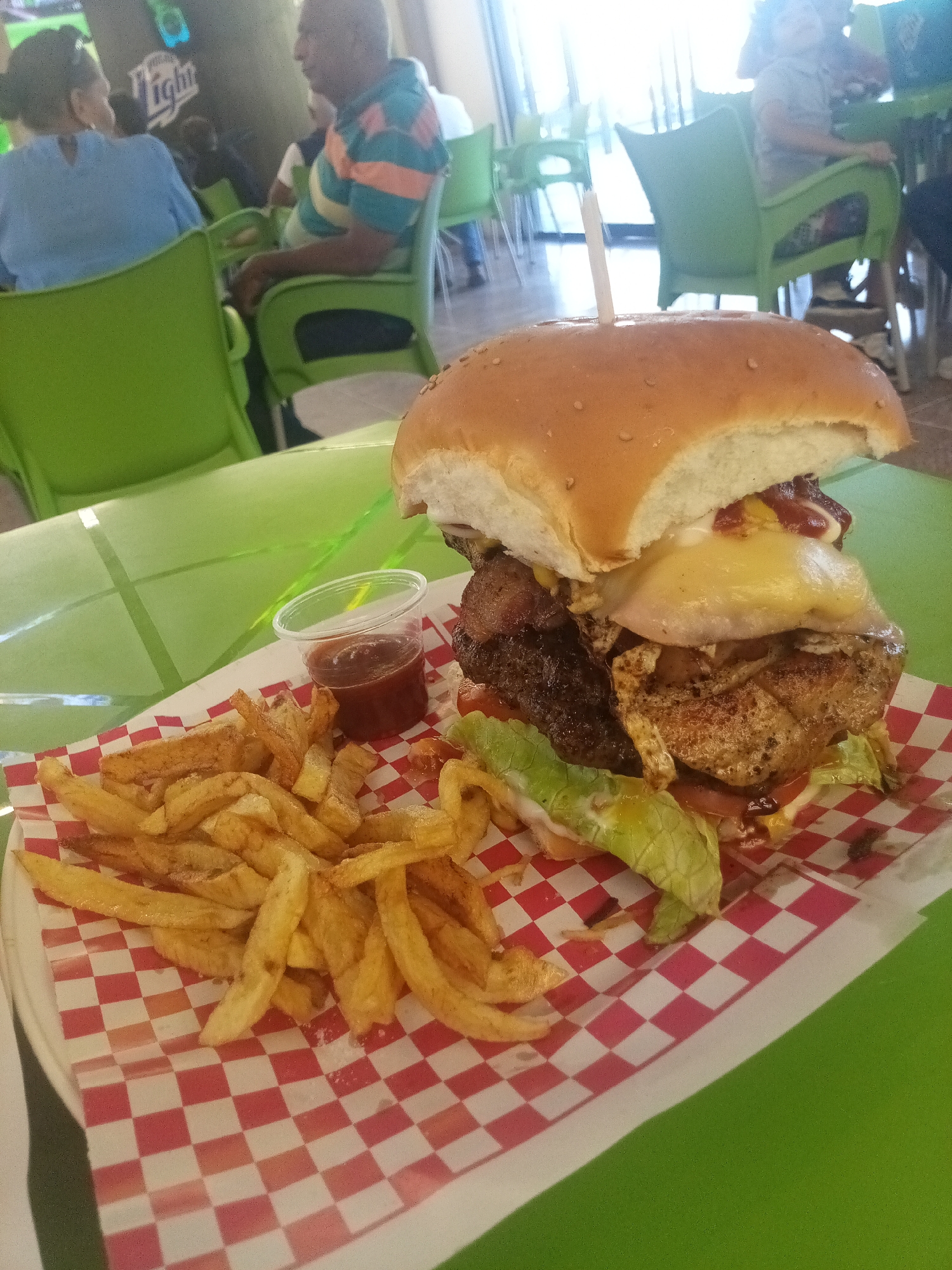 The image captures a close-up view of a substantial burger and fries meal placed on a table, likely in a restaurant or dining establishment. The burger is tall, featuring multiple layers that appear to include a beef patty, bacon, egg, cheese, and lettuce, held together by a bun pierced with a wooden skewer. A small plastic container holding a reddish sauce, possibly ketchup or barbecue sauce, sits beside the burger. The fries are plentiful and golden, arranged on a plate lined with a red and white checkered paper.
In the background, indistinct figures of people seated at tables are visible. The chairs are green, and there are bright green tabletops.
Based on the visual evidence and knowledge of the location, it is plausible that this image was taken at a restaurant or diner in Porlamar, Venezuela. However, without additional contextual information, it is impossible to confirm the precise location.