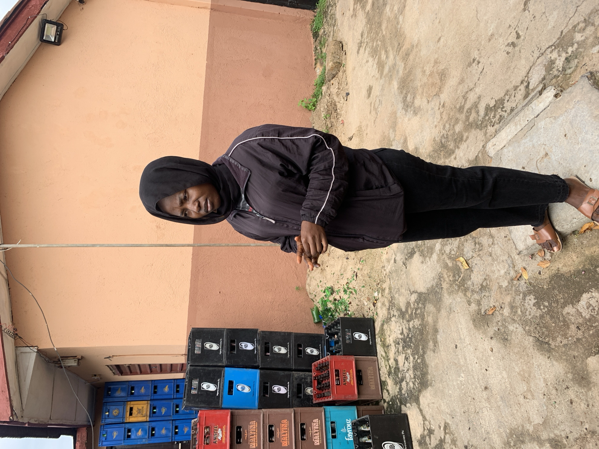 A medium shot captures a woman standing outdoors in a cluttered area. She wears a black hijab and a black jacket with white piping, paired with black pants and brown sandals. Her left hand is held up, fingers slightly apart, in a gesture that could indicate speaking or questioning. She is positioned to the right of a stack of colorful plastic crates, some blue and some yellow, which appear to be beverage crates. To the left of the woman and slightly behind her are numerous black and blue plastic crates, also filled with beverages, forming dense stacks. Among these crates, a red crate is visible, containing bottles. A pile of sand and broken green glass bottles is located to the right of the crates, indicating a possible waste disposal or previous accident. The ground is concrete and appears worn and damp. In the background, a light-colored building with a reddish trim is visible. The overall scene suggests an outdoor storage area or a small shop. The presence of beverage crates and bottles implies a commercial setting, possibly a distributor or a bar. The location is identified as Jos, Nigeria.