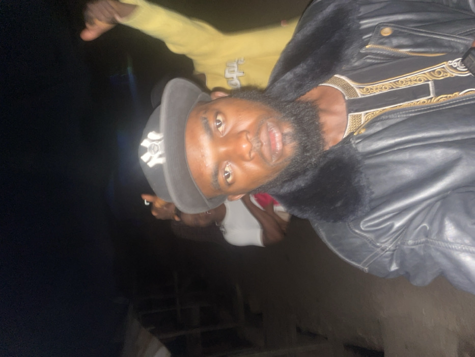 A dark and blurry image depicts a bearded man wearing a cap and a leather jacket with a fur collar. He also wears an embroidered garment. Another person, possibly wearing yellow clothing, is partially visible in the background, along with glimpses of other people. The picture's orientation is sideways, and the low lighting conditions make it difficult to discern details.