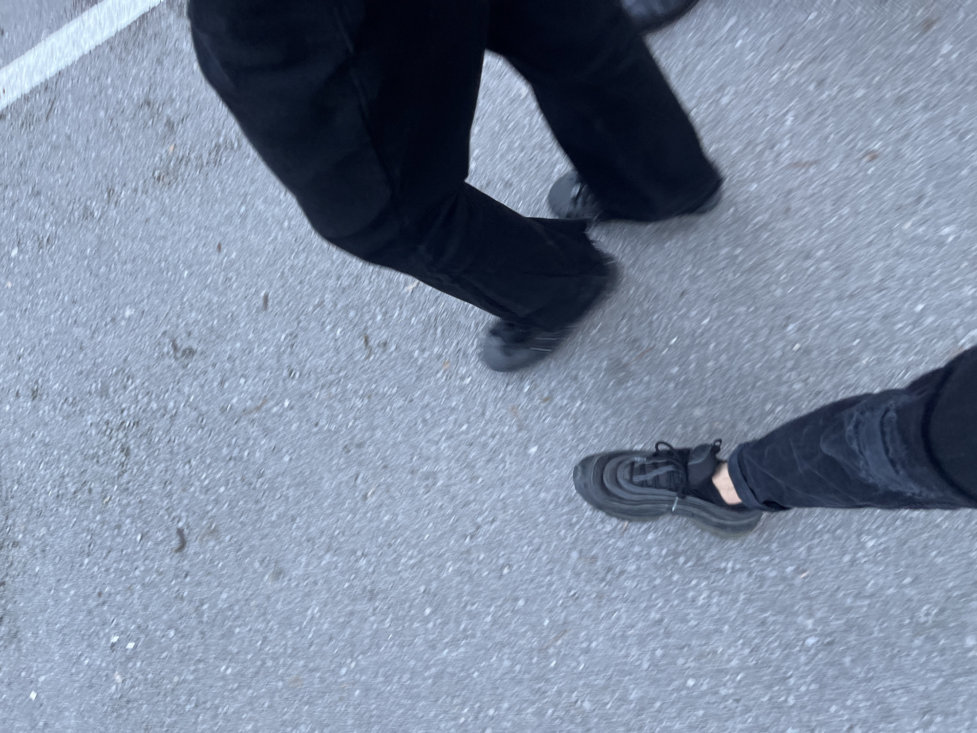 The image shows a ground-level view of two pairs of legs and feet walking on a gray asphalt surface. Both individuals are wearing dark-colored clothing, with one wearing black jeans or pants and dark sneakers. The other person is also wearing dark pants, and their shoes appear to be black, possibly boots or athletic shoes. The background is a blurred asphalt surface with no distinctive landmarks visible. The location is unknown.