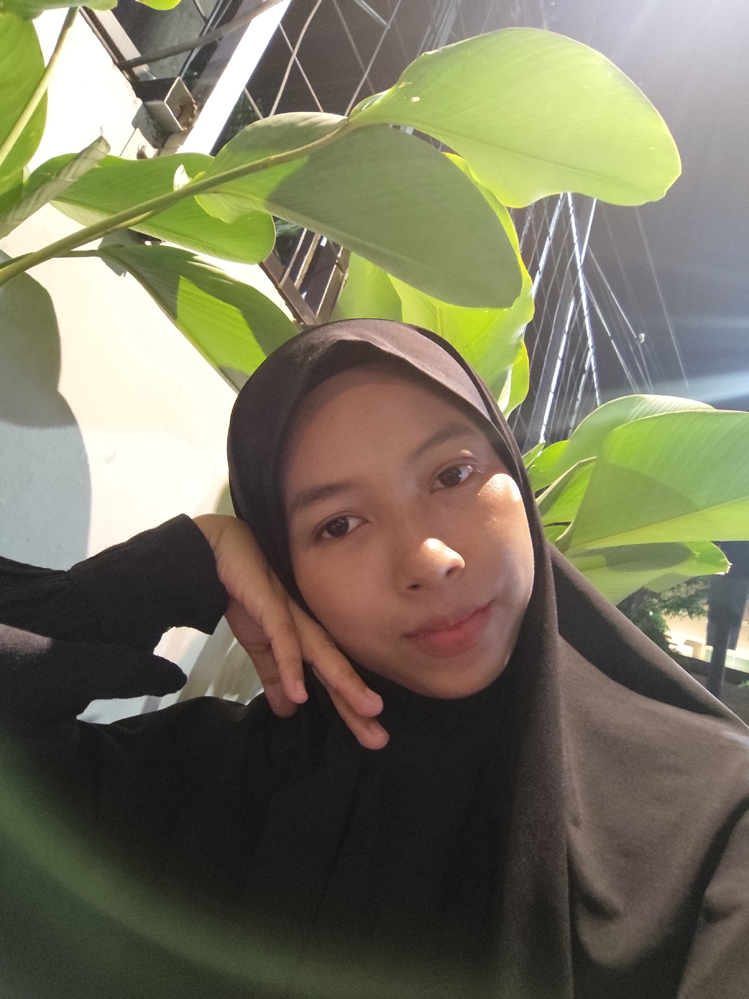 This is a selfie taken by a young woman wearing a black hijab and black clothing. She is resting her head on her hand, with her eyes looking towards the camera. Her lips are slightly parted, and she appears to be smiling subtly. In the background, there are large green leaves of a plant, and behind that, a blurry, out-of-focus background with what appears to be some metal structures and lights, suggesting an outdoor or semi-outdoor setting at night. The lighting suggests it is evening or night time. There is no visible text in the image.