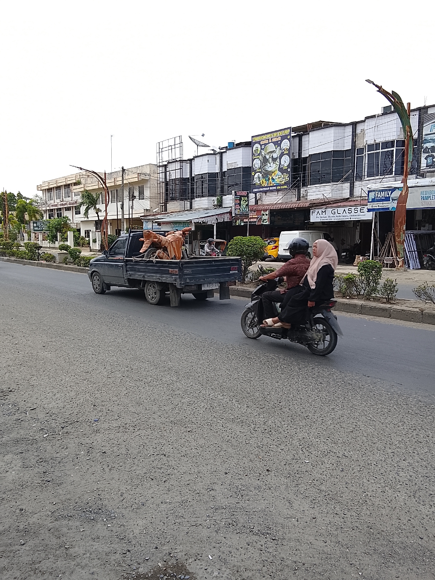 The photograph captures a street scene in Lhokseumawe, Indonesia. A black Isuzu pickup truck with license plate "BL 8375 ZT" transports what appears to be an arrangement of sculpted figures or decorative items in its bed. Moving in the same direction as the truck is a motorcycle with two occupants. The driver wears a helmet and a brown patterned shirt, while the passenger is wearing a hijab. Shops line the background, including "TOKO KUCING COPETLOVECAT'SHOP OPET LOVE CAT SHOP", "FAM GLASSES" and "FAMILY". The road surface shows signs of wear. Street lamps are visible along the roadside.