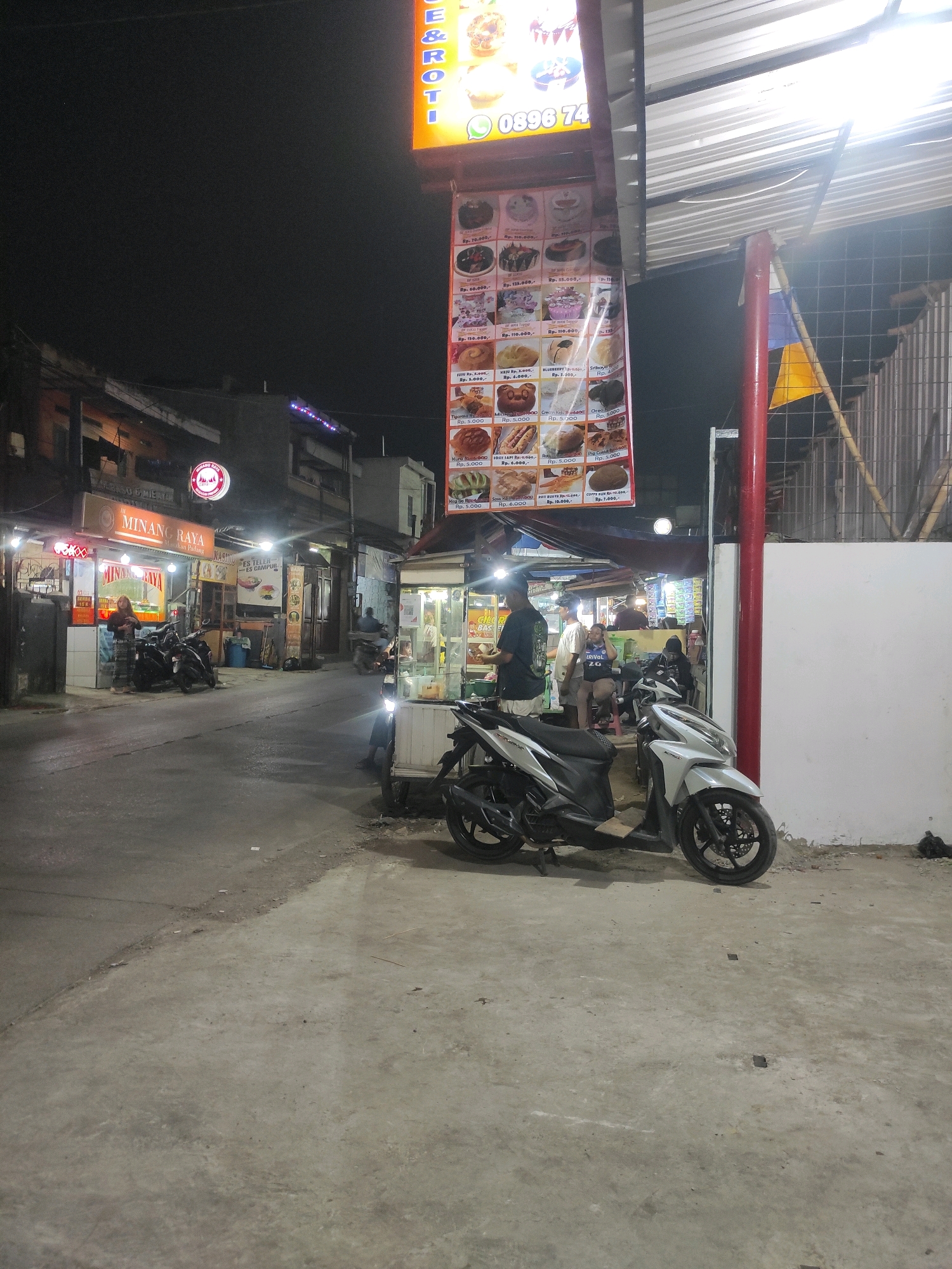 In Padalarang, Indonesia, an outdoor scene at night depicts a street lined with food stalls. A prominent sign, partially obscured, reads "MINE & Roti" in large letters and displays a phone number, "0896 74". Below this, a menu board shows various food items with prices, including options like "Roti Coklat" priced at Rp. 5.000 and "Kue Nastar" at Rp. 3.000.
Several motorbikes are parked on the side of the street, with two in the foreground, one white and one black and white. A few individuals are visible, some standing and some sitting near the stalls. One woman is standing at a stall labeled "MINANG RAYA" with a red circular logo. Another stall appears to be selling drinks. The street is paved, and the surrounding buildings are dimly lit, with some lights from the stalls illuminating the area. The overall atmosphere suggests a lively night market or street food scene.