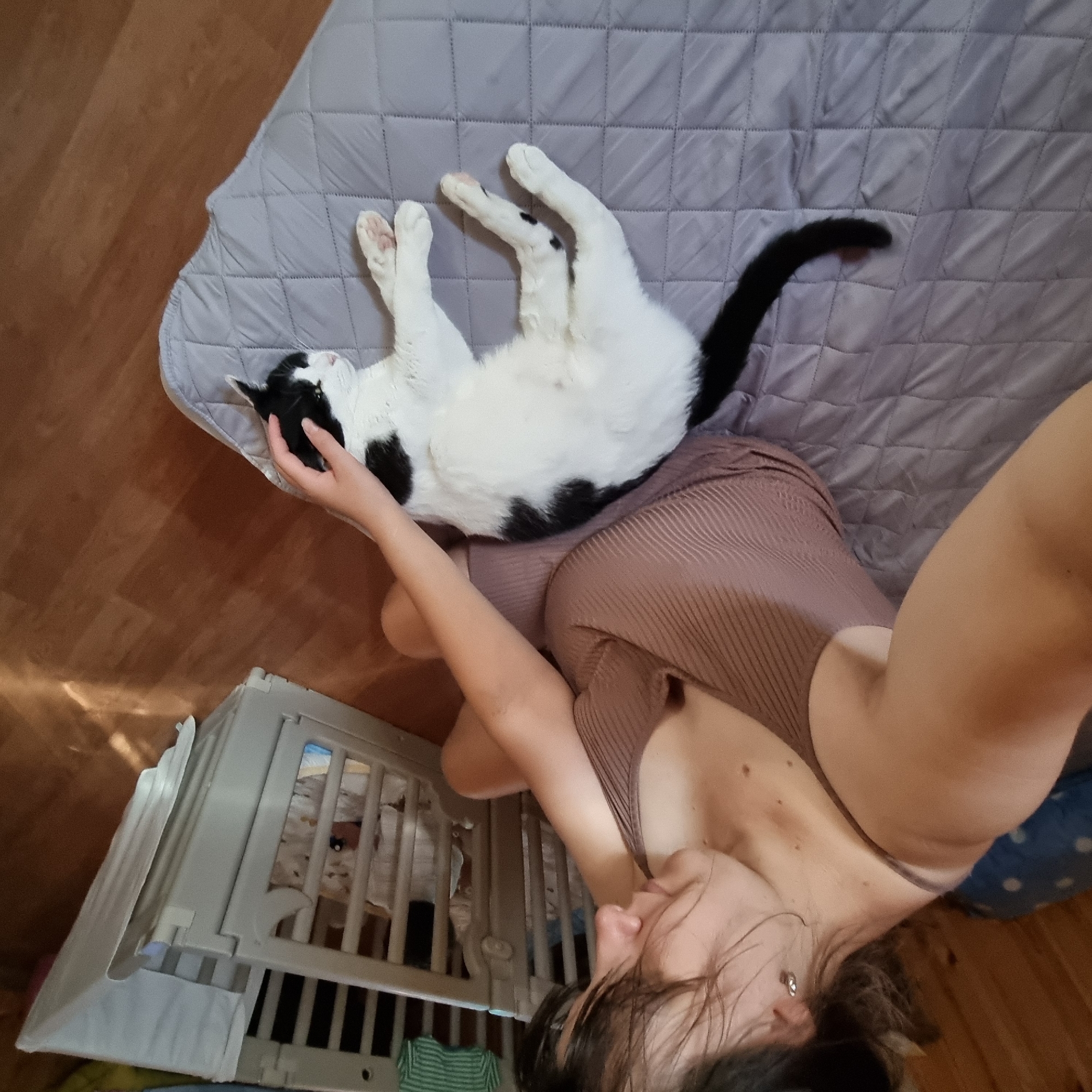 A person with dark hair is sitting on a gray quilted surface, petting a black and white cat. The person is wearing a brown ribbed tank top. The cat is lying down on its side, with its head resting on the person's hand. In the background, a light-colored baby crib is visible. The floor appears to be made of wood. The overall setting suggests an indoor domestic environment. There is no information to suggest this scene is located in Dunnae-myeon, South Korea.