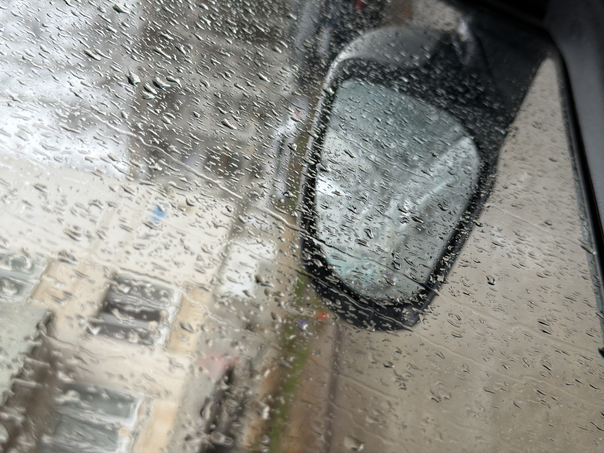 The image depicts a view through a rain-streaked car window. A side mirror is visible, also covered in rain. Outside the window, blurred shapes suggest buildings and possibly a sidewalk. While I cannot confirm the specific city of Chaikovsky, Russia, the scene appears to be an urban environment.
