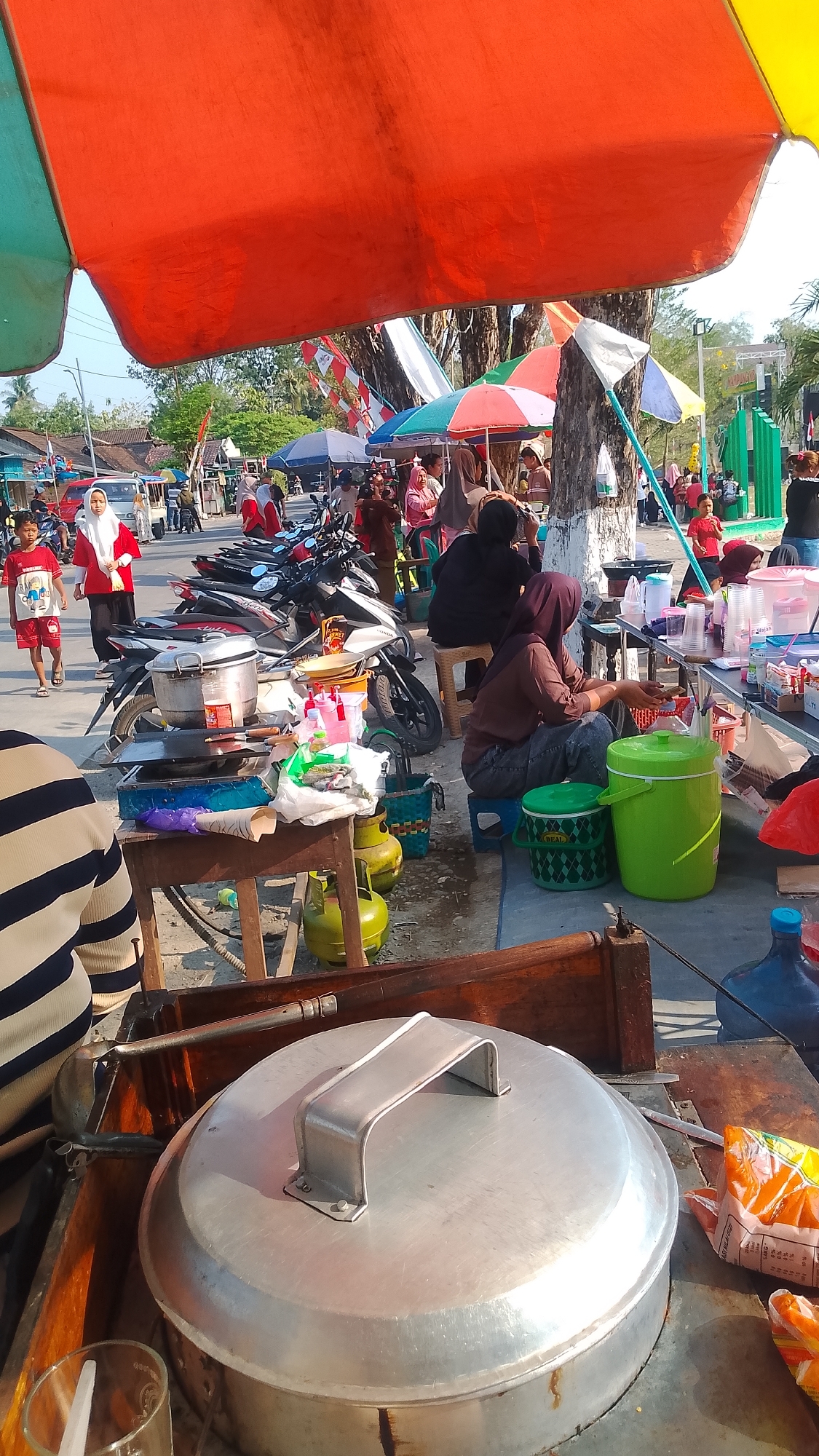 This is an outdoor scene in Karanglangu, Indonesia, likely a market or festival, under a large orange and green awning. In the foreground, a large, round, silver pot with a metallic handle sits on a wooden cart. To its left is a glass with a white straw. To the right of the pot, part of a plastic bag with orange and white packaging is visible. Behind the cart, two green propane tanks are propped up. Several motorcycles are parked in the middle ground. A young boy in a red t-shirt and red shorts walks past the motorcycles. Further back, several women are seated at a table under a colorful umbrella, some of them wearing hijabs. They appear to be preparing or selling food or drinks. In the background, more stalls, people, and trees are visible. The lighting suggests it is daytime, likely late afternoon due to the long shadows.