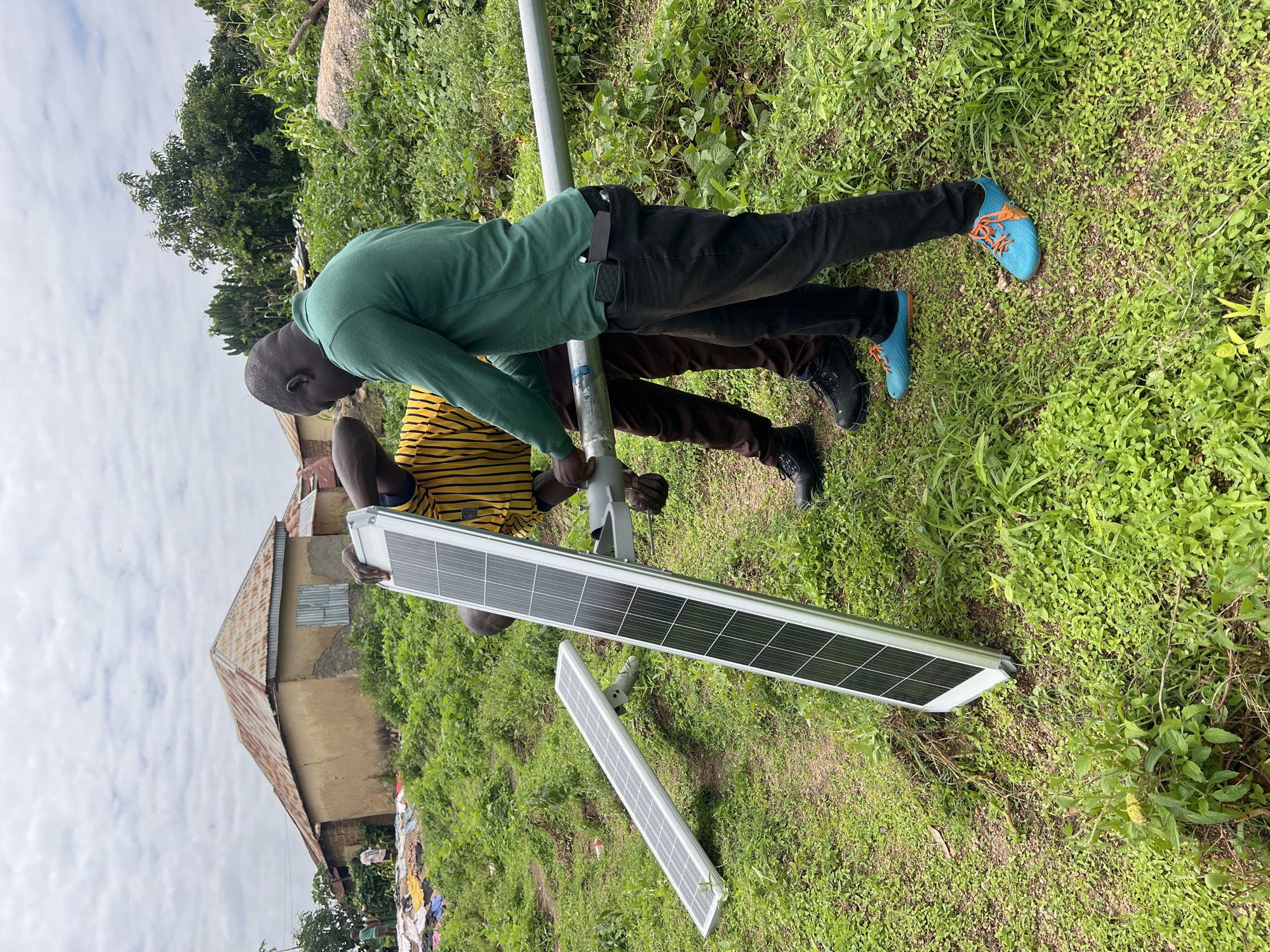 Two men are in a grassy area, likely in Dong, Nigeria, working on installing solar panels. One man, wearing a green long-sleeved shirt and black pants, is holding a metal pole. The other man, in a yellow and black striped shirt and brown pants, is assisting him. Two solar panels are visible on the ground, one leaning vertically and the other lying horizontally. In the background, there are traditional Nigerian buildings with corrugated metal roofs and dense green vegetation. The sky is overcast.
