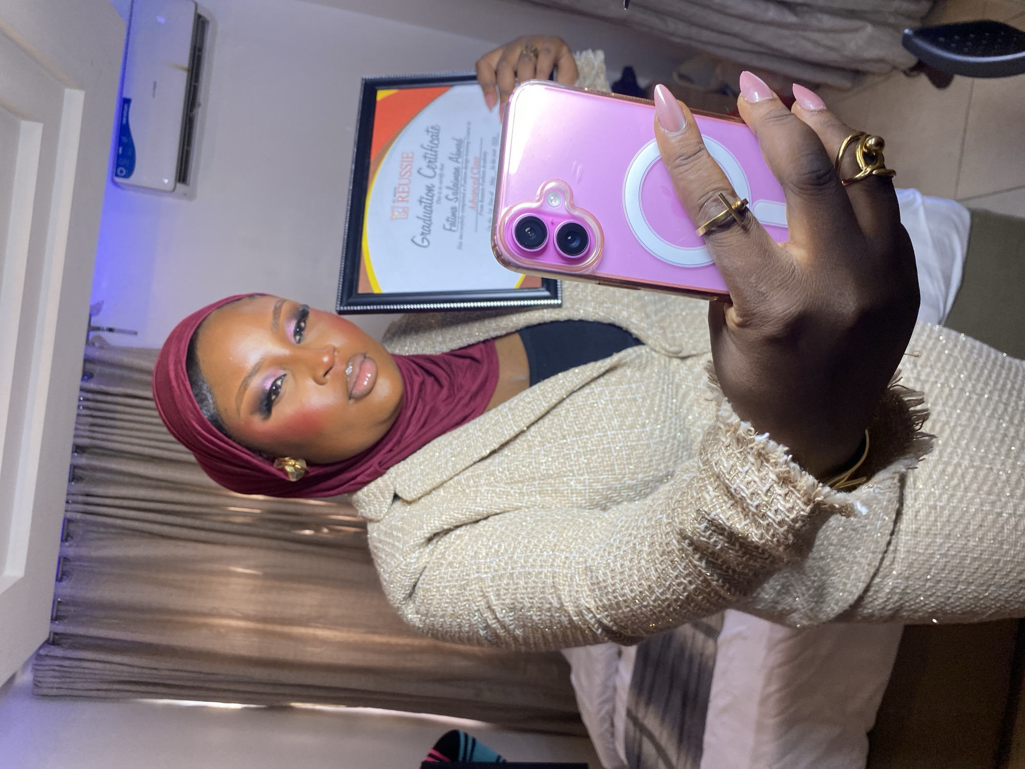 A woman wearing a burgundy hijab and a light-colored, tweed-like jacket is taking a selfie while holding up a framed graduation certificate. The certificate appears to be from "REUSSIE" and mentions "Graduation Certificate" for "Fatima Suleiman Ahmed" in "Advanced Class" from "Reussie Fashion Academy." The woman is using a pink phone with a visible magnetic ring on the back. The setting appears to be indoors. The location is Gura Top, Nigeria.