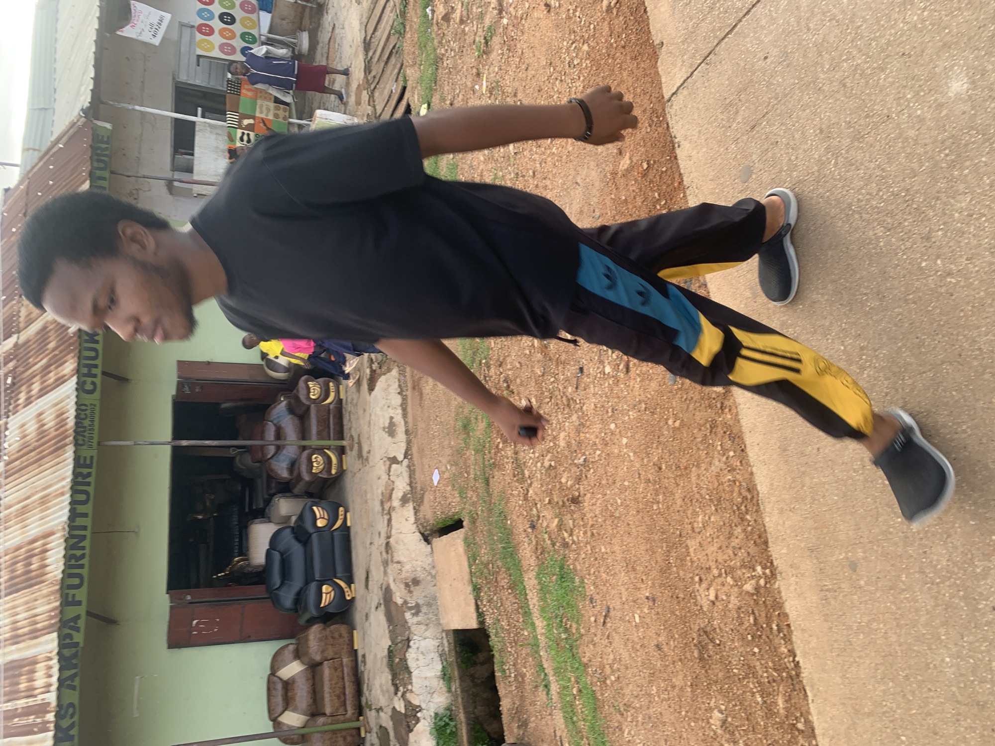 The image shows a young Black man standing outside a shop. He is wearing a black t-shirt and black and yellow athletic pants. He has a bracelet on his left wrist. He appears to be looking down at his feet. The shop behind him has a sign that reads "AKS AKPA FURNITURE CHUK." The shop is filled with various chairs and sofas. There are other people in the background, including a person in a blue jacket and red shorts. The ground is a mix of dirt and concrete. The overall scene appears to be a street in Jos, Nigeria, during daylight hours. The weather appears to be warm and dry.