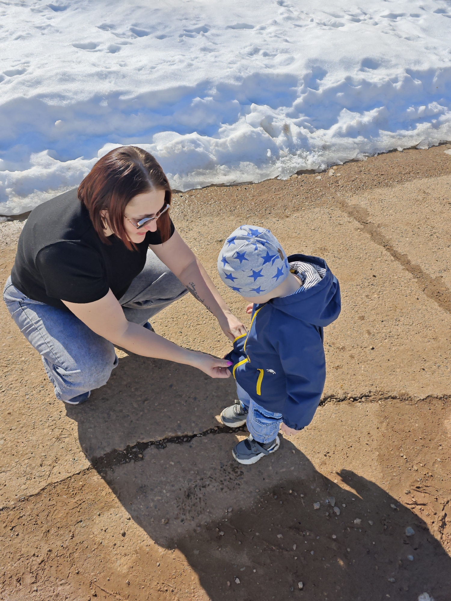 A woman is crouching and interacting with a small child in an outdoor setting. The woman has reddish-brown hair, is wearing sunglasses, a black t-shirt, and light blue jeans. The child wears a blue jacket with yellow trim and a light gray hat with blue stars. The woman appears to be adjusting or interacting with the child's jacket.
The ground surface is a cracked, concrete-like material with some patches of brown soil. In the background, there is a snow-covered area. The location is likely in an area with both snow and exposed ground, suggesting it is a place where snow is melting or is not entirely covering the ground. The city is Марковский, Russia.
