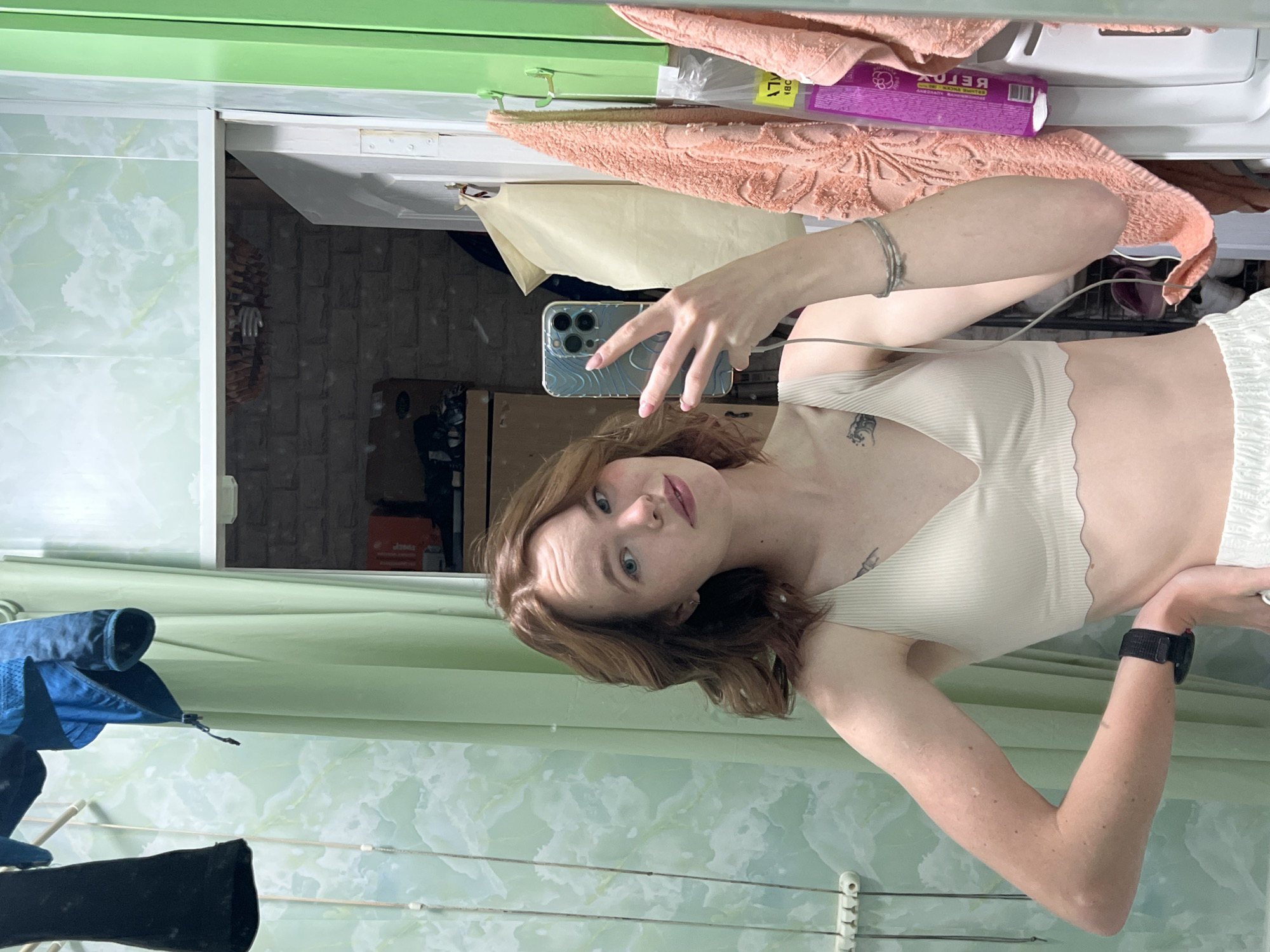 A person is taking a selfie in a bathroom. The person is fair-skinned with reddish-brown hair and blue eyes, wearing a light-colored top and a watch on their left wrist. They are holding a smartphone in their right hand, presumably to take the picture. The bathroom has a green and white marble patterned wall. There's a clothes drying rack with clothing hanging on it in the lower left corner. A cabinet with a mirror is visible above and behind the person. Towels and a box of "BELOX" (spelled in Cyrillic) are on a shelf inside the cabinet. The reflection shows a brick wall and items inside the cabinet, including what appears to be a cardboard box. The location is Chaikovsky, Russia.