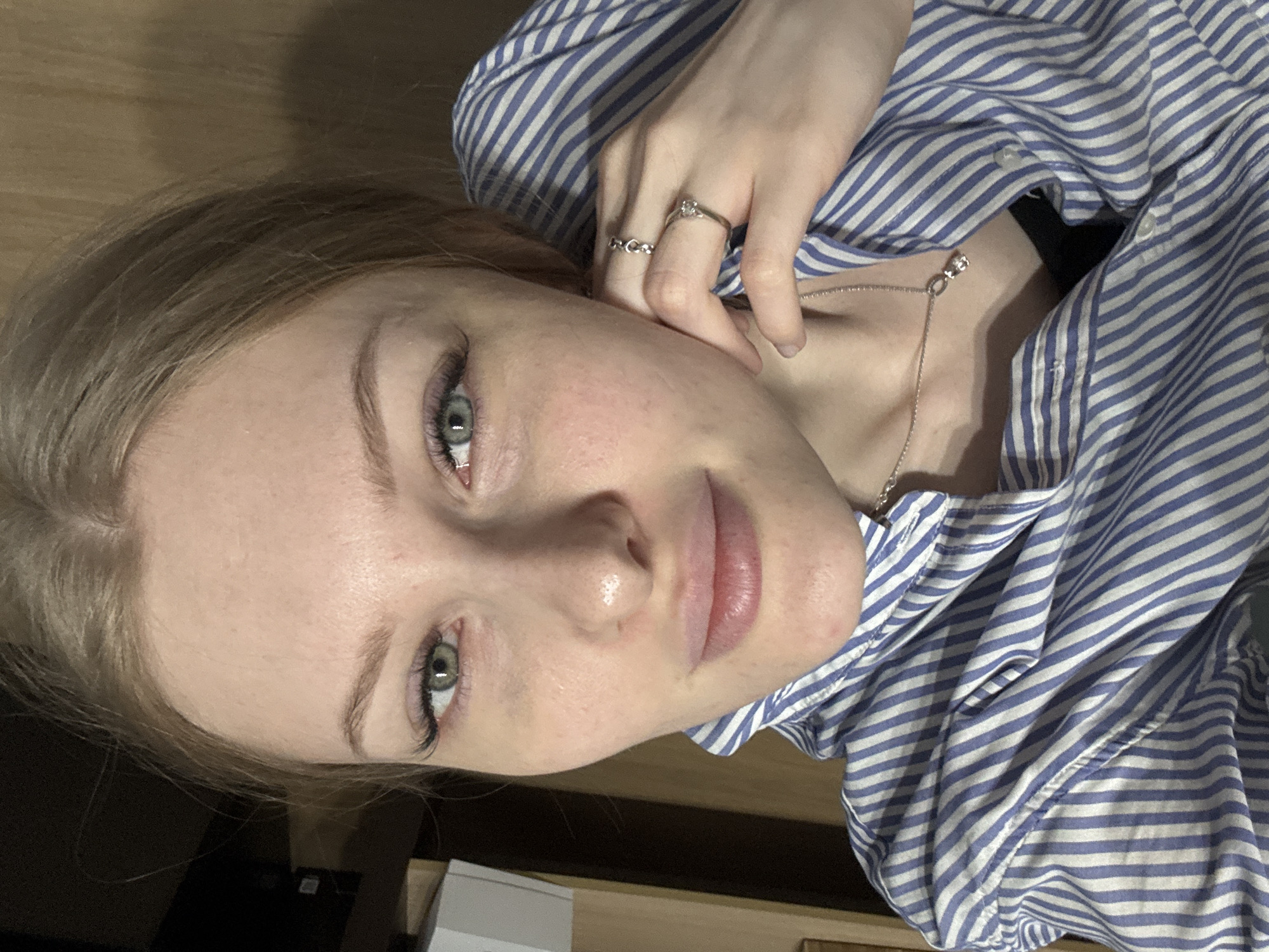 A close-up, slightly angled portrait of a young woman. She has light blonde hair, fair skin, and green eyes. She is wearing a blue and white striped shirt. Her hand, adorned with two rings, is near her face. A delicate necklace is visible around her neck. The background appears to be an indoor setting with wooden surfaces. The location (Perm, Russia) is not discernible from the image.