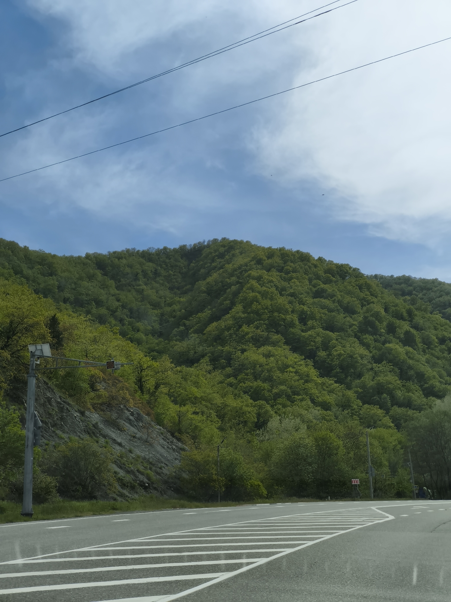 A road intersection in Pshada, Russia, with a mountain densely covered in green trees in the background. An overhead power line stretches across the sky, which is blue with scattered clouds. Road markings are visible, indicating a turn in the road. A utility pole with a traffic control device is to the left. A signpost with chevron markings is visible on the right side of the road.