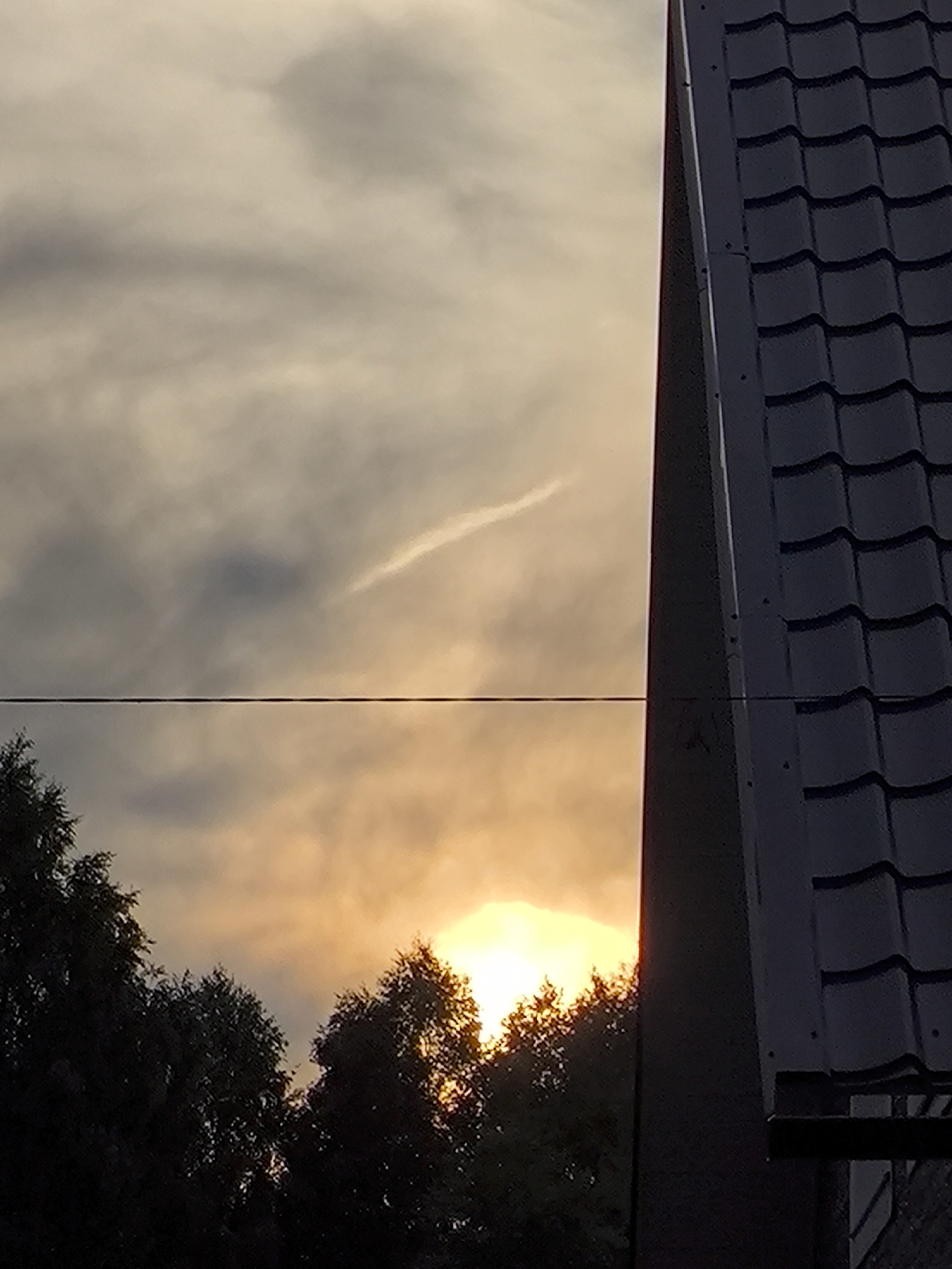 A low-angle, full shot captures a sunset scene in an urban environment. The sky is a gradient of warm yellows and oranges, with scattered gray clouds. A thin, wispy cloud streak is visible in the upper portion of the sky. In the foreground, a dark, grey tiled roof dominates the right side of the frame, with a portion of a dark grey wall or building facade extending from its left edge. A thin, black horizontal line, possibly a power line or wire, bisects the image horizontally. Silhouetted against the bright sky, a dense cluster of dark green trees occupies the lower left portion of the frame. The overall impression is one of twilight in a built-up area.