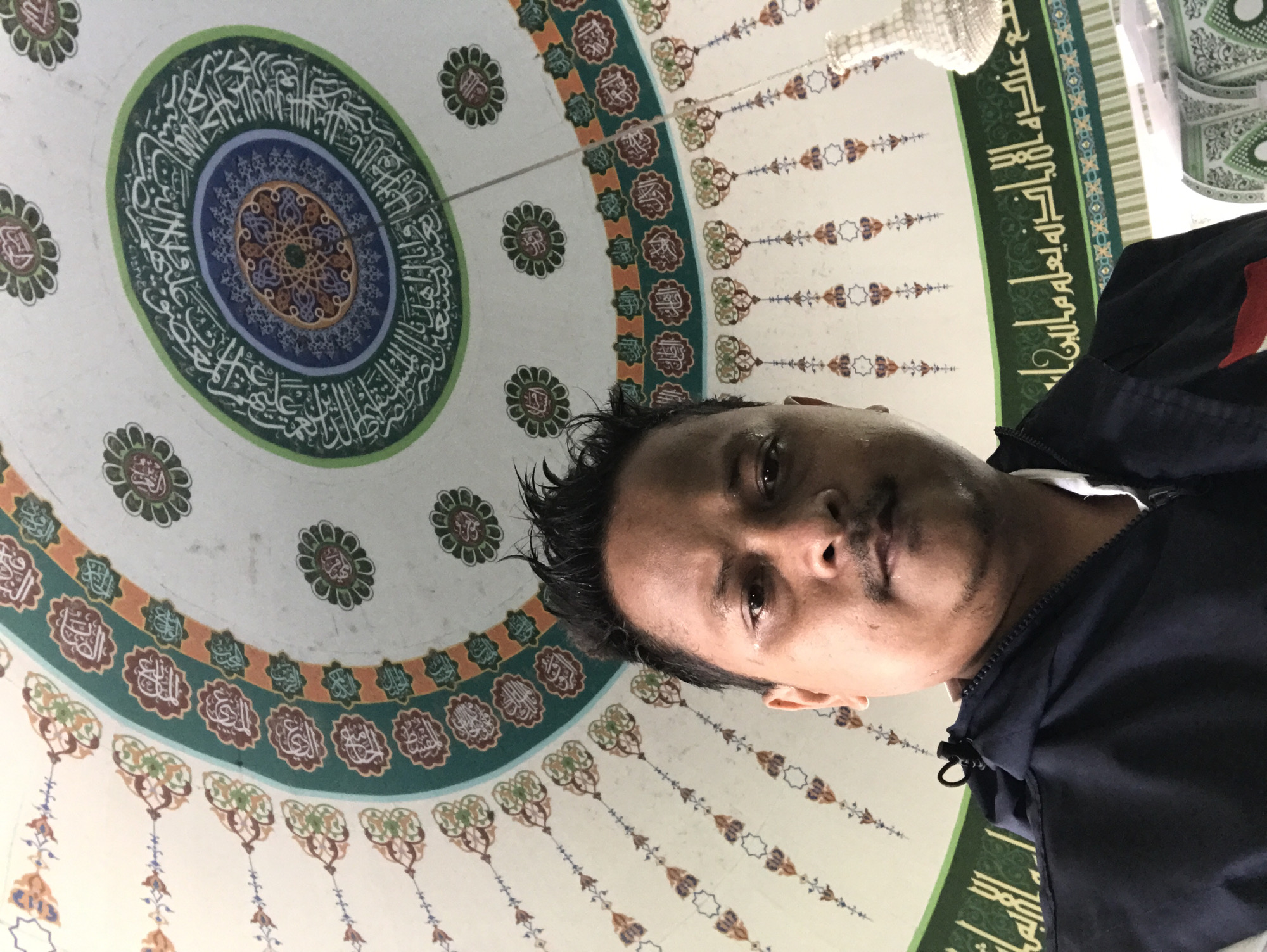 A man is positioned in the foreground, appearing to be taking a selfie or being photographed from a low angle. He has dark hair and brown skin, wearing a dark blue jacket with a white collar underneath. The background reveals an elaborately decorated ceiling, characteristic of Islamic architecture, potentially inside a mosque. This ceiling exhibits a large circular design with concentric layers of ornamentation, featuring geometric patterns, floral motifs, and Arabic calligraphy in hues of blue, green, brown, and white. Additional details include hanging decorations and green panels with further inscriptions along the edge of the ceiling. The image's origin is Jantho, Indonesia.