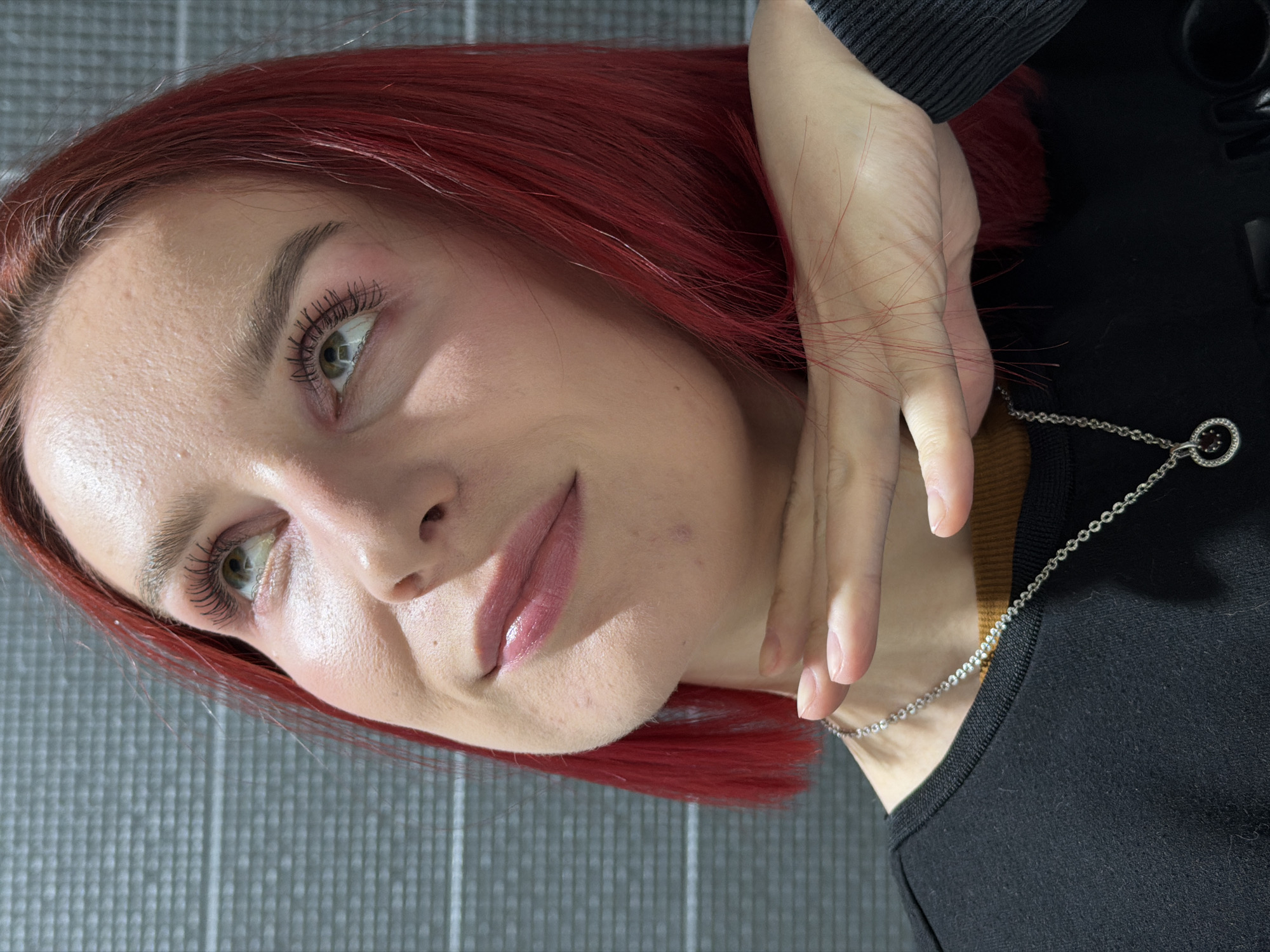 This is a medium close-up, angled portrait of a young woman, rotated 90 degrees clockwise. The primary subject is a woman with striking, vibrant red hair, which appears to be dyed. Her hair frames her face and some loose strands are visible, particularly intertwined with her hand. She has light-colored eyes, appearing green or hazel, and looks upwards and slightly to the left, conveying a calm and thoughtful expression, possibly with a subtle, gentle smile. Her face shows light makeup, including well-defined eyebrows, mascara on her eyelashes, and glossy pink lip gloss.
She is wearing a dark, likely black, long-sleeved top with a ribbed cuff, and a brown or tan collared garment underneath. A silver chain necklace with a circular pendant, embellished with small dark red or black stones, is visible around her neck. Her left hand is gently placed near her jawline and neck. The background is a uniform grey surface with a subtle grid or textured pattern, suggesting an indoor or studio environment. The lighting is soft and even, highlighting her features. No specific activity is depicted beyond posing for the photograph. There is no visible text in the image. The location context is Chaikovsky, Russia.