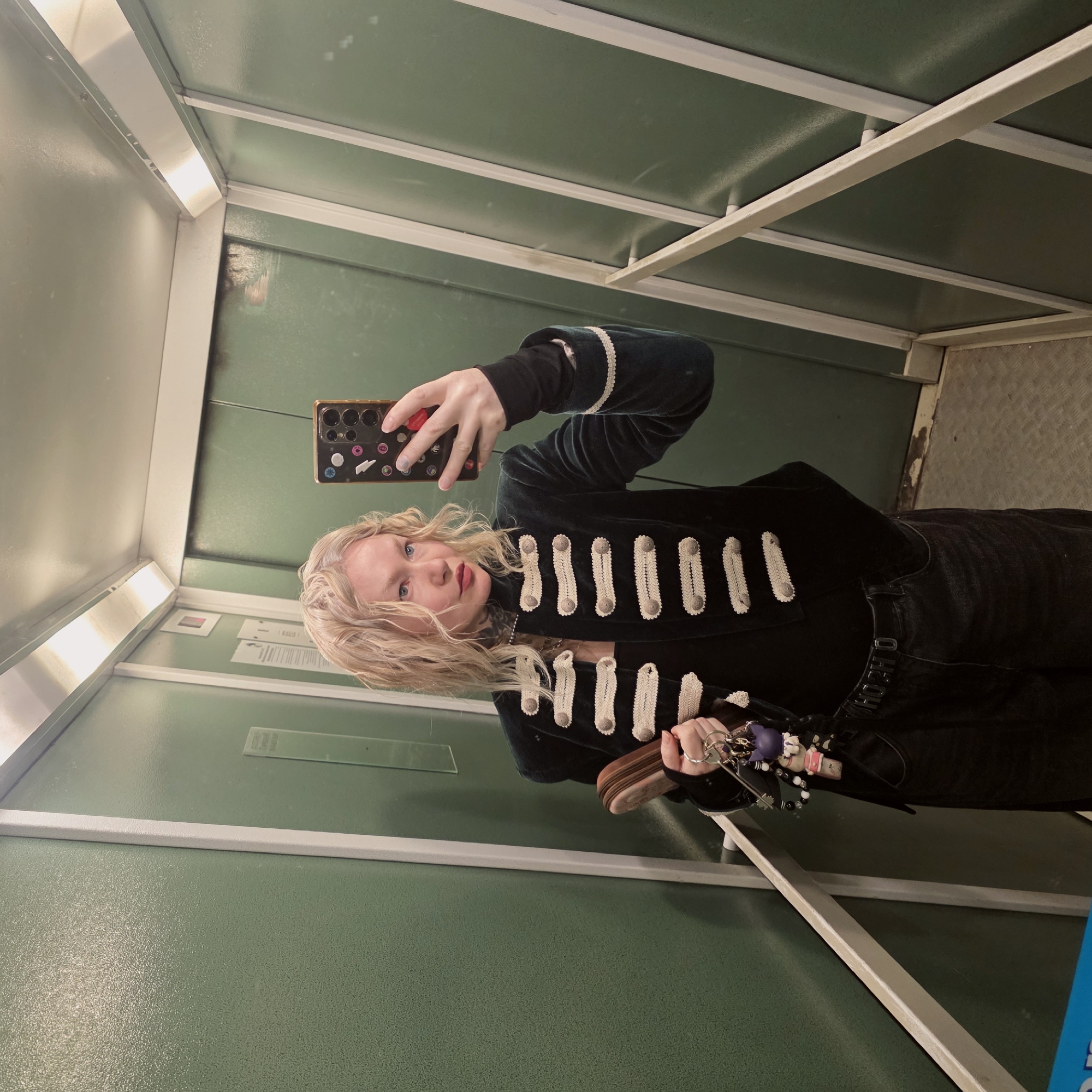A woman is taking a selfie in what appears to be an old, possibly Eastern European elevator. The elevator has green paneled walls, with white trim. The woman is wearing a black velvet jacket with white decorative trim, black pants, and a belt with a silver buckle. She is holding a brown wallet and a set of keys. The location is possibly Saint Petersburg, Russia. The setting is an old elevator.