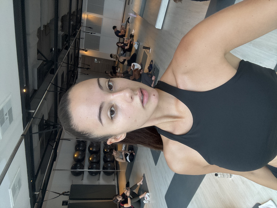 A female subject is depicted in the foreground, capturing a selfie within a modern fitness or yoga studio. She has dark hair pulled back into a high ponytail and is wearing a black high-neck athletic top. Her gaze is directed at the camera with a neutral expression, lips slightly parted, and a small, distinct mole is visible superior to her upper lip.
In the background, approximately ten to twelve other individuals are observed, seated or resting on individual dark grey exercise mats spread across a light-toned floor. These individuals are dressed in athletic attire; several are actively engaged with personal electronic devices, including mobile phones and laptops.
The studio environment features an exposed, industrial-style ceiling with visible dark structural beams, conduits, and ventilation systems. Track lighting fixtures are installed overhead. Along the left wall, a multi-tiered rack holds several black exercise balls, and a vertical silver pole is positioned adjacent. Multiple black suspension training straps are affixed to the ceiling structure. Additional items on the floor include clear water bottles and white hand towels.
The setting is a fitness studio located in Bangkok, Thailand; however, the specific address or detailed location within the city is not discernible from the visual information provided in the image.