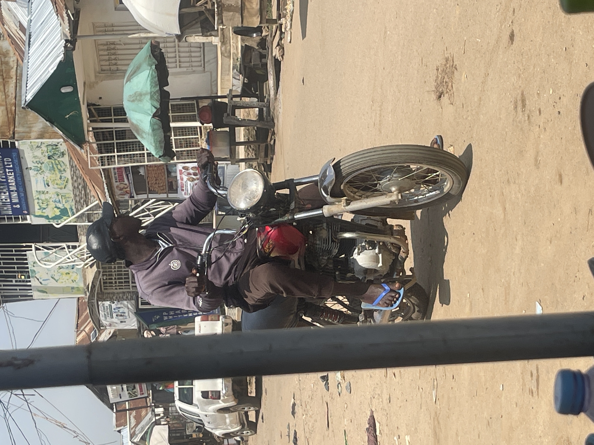 The image depicts a street scene in Abuja, Nigeria, under a bright sky. In the foreground, a person wearing a dark cap, a purple hooded jacket, and brown trousers is riding a motorcycle. The person's left foot is visible, wearing a blue flip-flop. The motorcycle itself is dark with a red fuel tank. Behind the motorcycle, a white pickup truck is parked on the dirt road, which is littered with debris. The background shows various buildings with corrugated metal roofs and awnings, suggesting a commercial or residential area. A green umbrella is open, and a white umbrella is partially visible to the right. Signs with text, possibly advertisements or shop names, are displayed on some buildings. The overall impression is one of a bustling, informal urban environment.