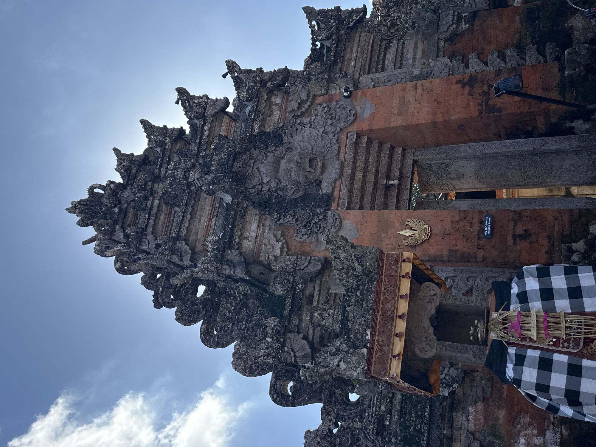 The image captures a detail of an ornate, Balinese temple gate, likely in Ubud, Indonesia. The architecture is characterized by intricate stone carvings, featuring mythological figures and floral motifs, set against a backdrop of red brick. The style suggests a traditional Hindu temple. The sun is bright, indicating it is likely daytime, and the sky is a clear blue with a few white clouds, suggesting fair weather.
In the foreground, to the right, is a partially visible offering or altar adorned with a black and white checkered cloth, a common sight at Balinese temples. A small, elaborately decorated structure is also visible, perhaps part of the temple's ritualistic setup. The sunlight glints off some of the stone elements and creates a bright halo effect around the upper parts of the temple structure, suggesting the sun is positioned behind it.
The overall impression is one of cultural richness and spiritual significance, typical of the religious sites found in Bali. There is a small sign visible on the temple wall with text, partially obscured, but it appears to be an instruction or information for visitors, possibly relating to behavior within the temple grounds. The image is oriented vertically, with the temple structure dominating the frame and extending upwards towards the sky.