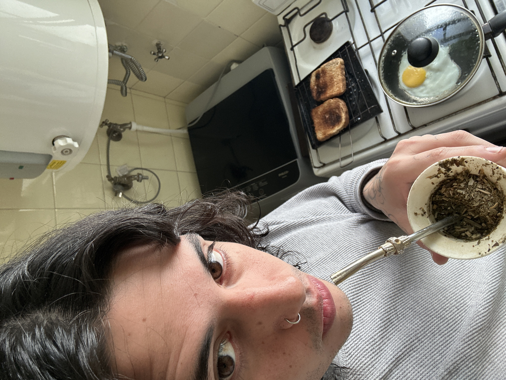 The photograph is a selfie of a person with long dark hair, brown eyes, and a nose ring. The person is wearing a gray sweater and is holding a mate gourd with a bombilla to their lips.
In the background, there is a kitchen scene. A white water heater is mounted on a tiled wall. To the right, there is a microwave and a white stove with two slices of toast on a grill and a fried egg cooking in a pan.
The setting appears to be a modest kitchen, possibly in a residence. While the image provides some visual clues, there is no way to determine with certainty where this is without additional evidence.