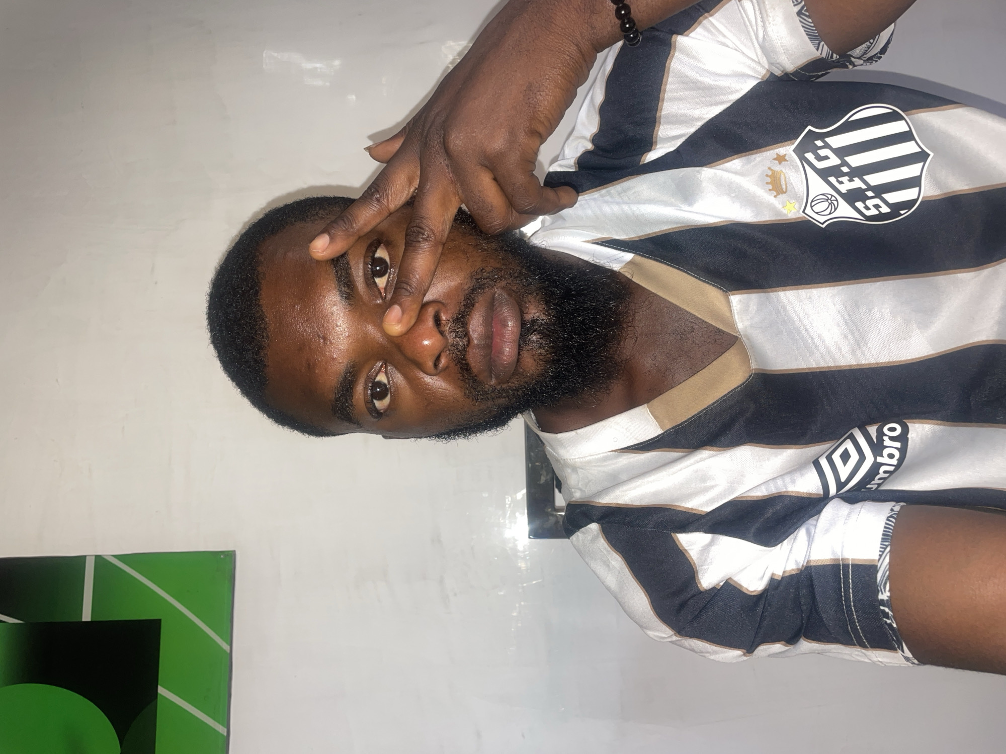 An image shows a dark-skinned man with a beard. The man is wearing a striped white and black soccer jersey with a logo on the upper right shoulder area. He is holding his fingers to his face, covering one eye. There is a green object at the bottom left of the frame. The location of the image is not discernible as being within the city of Jos, Nigeria.