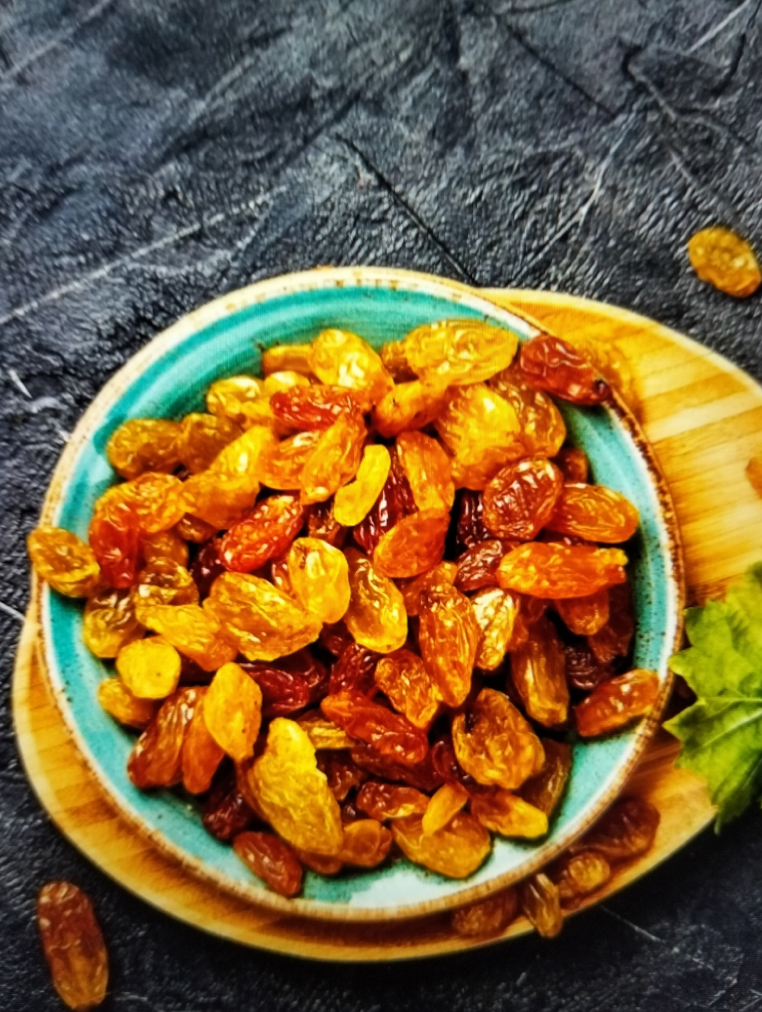 A high-angle, close-up shot features a teal-rimmed bowl filled with a generous portion of golden and dark brown raisins. The bowl rests on a rustic wooden platter, which in turn is set against a dark, textured surface, possibly slate or concrete. A few stray raisins are scattered on the dark surface, one to the left of the bowl and another in the upper right corner. A hint of green leaf is visible on the right side of the platter. The lighting is soft, creating subtle shadows and highlighting the plump, shriveled texture of the raisins. There are no people or animals present. The scene is static and appears to be a food still life. The image does not contain any elements that can be used to identify a specific location in Payagaboh, Indonesia.