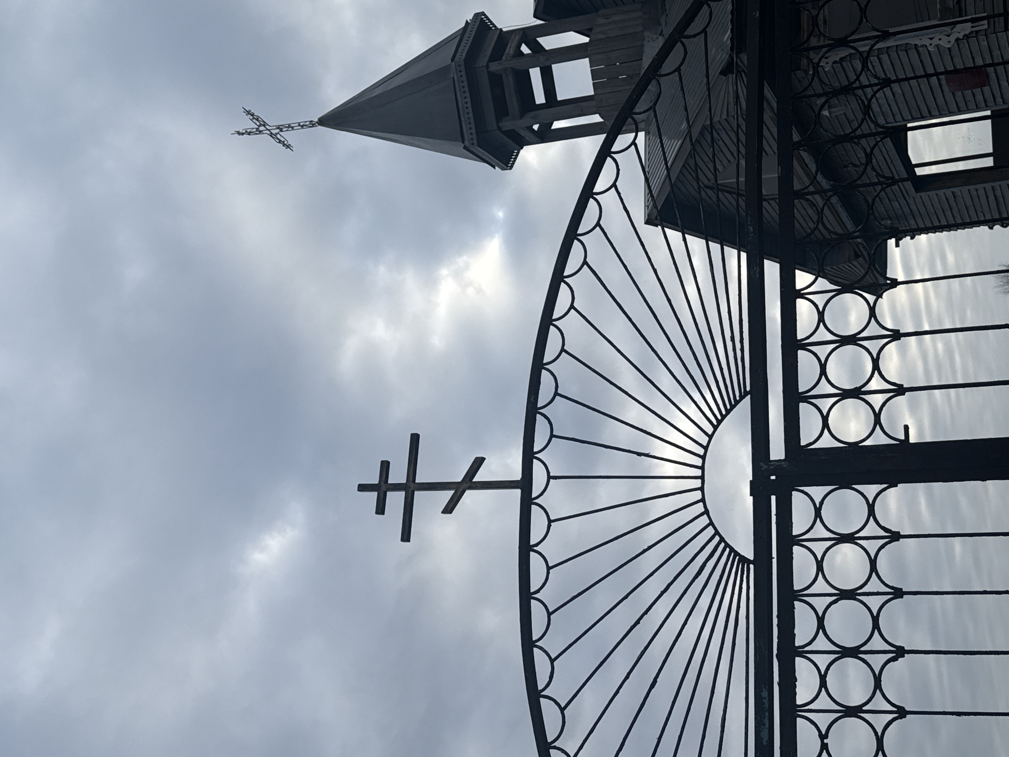 The image shows an upward view of a wrought-iron gate featuring a cross, with a Russian Orthodox church tower visible in the background. The church has a slender, multi-tiered structure topped with another cross. The sky is overcast, providing diffused light. The gate has a radial design in the upper portion, leading to a series of circular patterns in the lower part. The church appears to be located in Фоки, Russia.