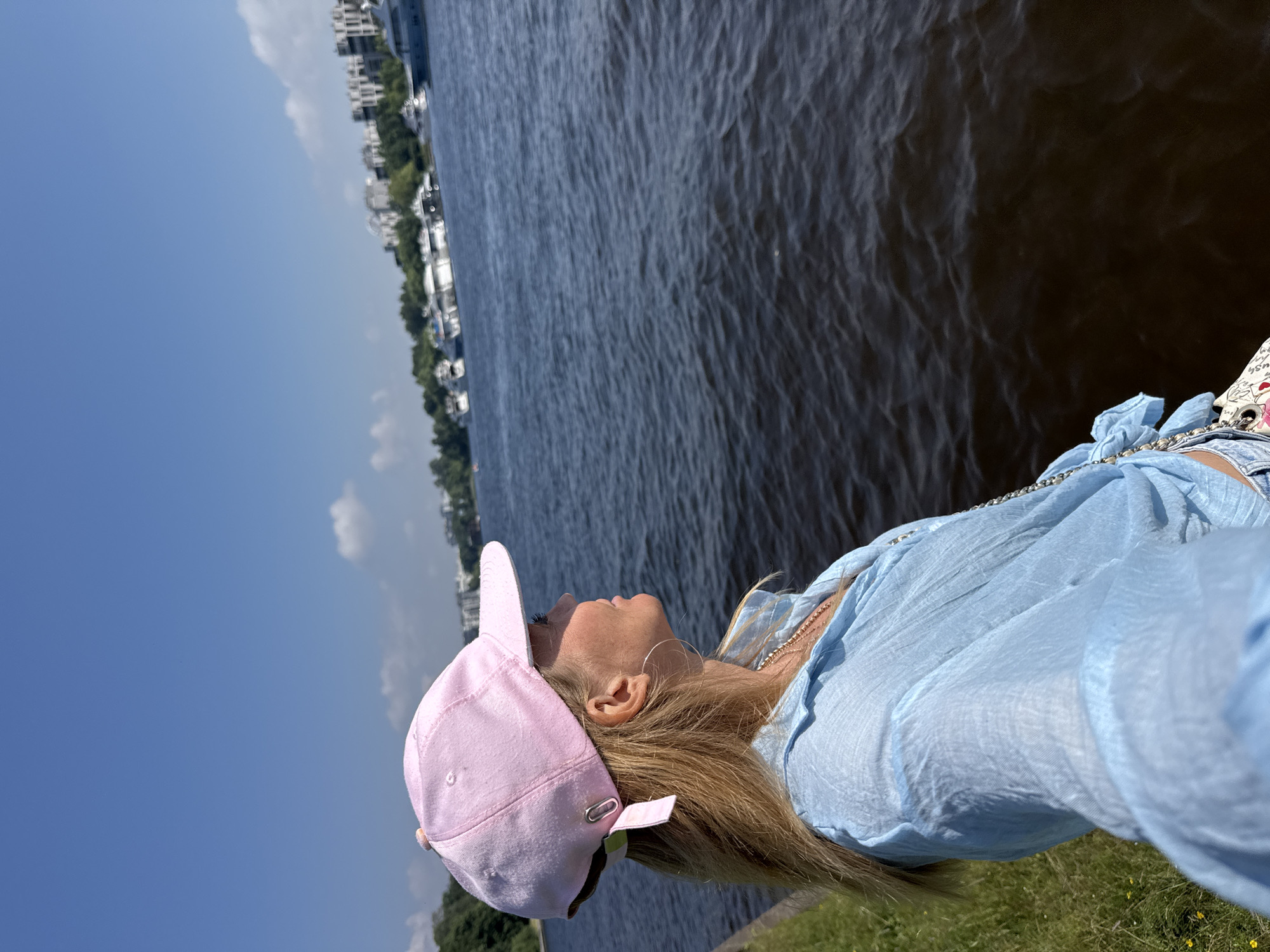 A blonde woman wearing a pink baseball cap and a light blue, sheer, long-sleeved shirt is captured in a selfie. Her head is tilted upwards, looking towards the sky. She has blonde hair and is wearing a silver hoop earring. The woman appears relaxed and content.
The setting is outdoors, beside a body of water that appears to be a river or bay. In the background, across the water, is a cityscape with white buildings, trees, and some boats docked. The sky is clear and blue with a few wispy white clouds. The sun is shining, suggesting it is daytime, likely midday given the brightness. The ground in the foreground is covered with green grass and some yellow flowers.
The image is oriented vertically, with the woman on the right side of the frame. The overall mood is peaceful and summery, with the woman enjoying the scenery and the weather. There is no visible text in the image. The location context indicates this is Saint Petersburg, Russia, and the cityscape in the background is consistent with this location.