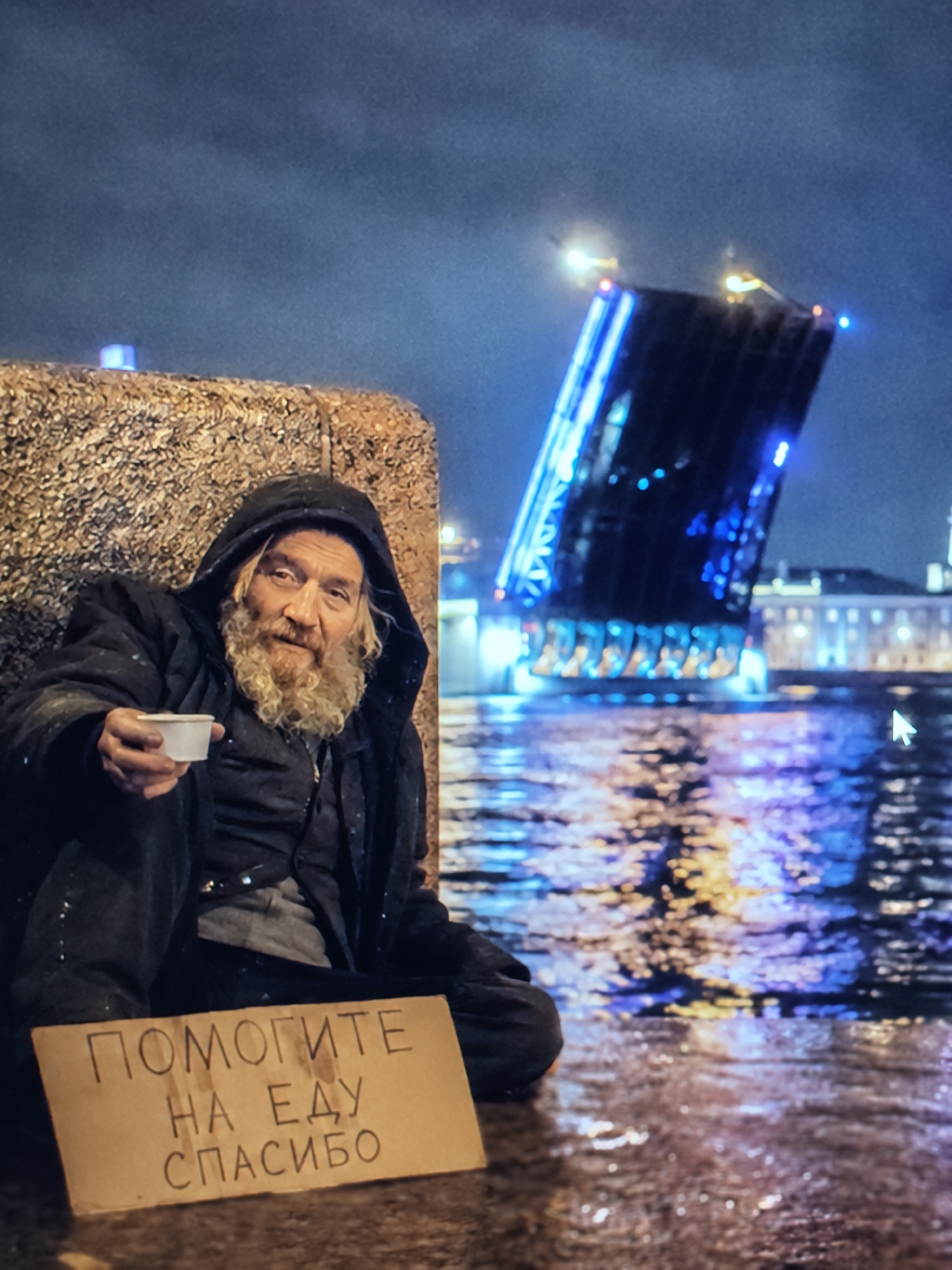 A man sits on the ground in front of a textured, golden-brown structure, holding a white cup in his outstretched right hand. He wears a dark, hooded jacket and has a long, light-brown beard and wavy hair. A cardboard sign rests on the ground in front of him with Russian text. The background shows a body of water with colorful reflections of lights from a large, illuminated bridge that is raised in the center. Buildings line the far bank of the water. The scene appears to be at night.