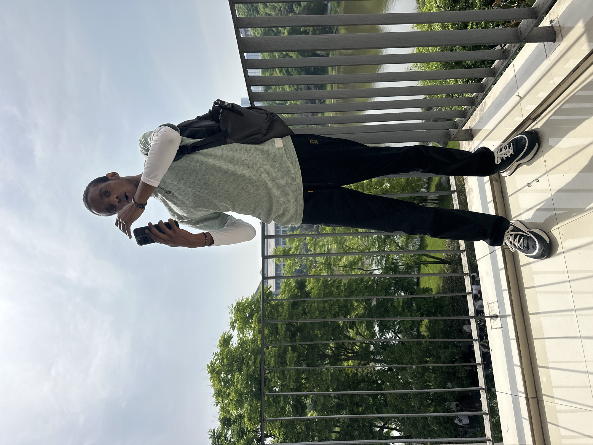 A man stands on a balcony with a metal railing overlooking a park or garden. He is holding a phone in his hands and appears to be taking a picture. He is wearing a light green sweater, black pants, and black and white sneakers. He also has a black backpack on his shoulders. The balcony has a tiled floor. Beyond the railing, there are trees and what appears to be a body of water.
The information about the location of this image in Jiangjin, China is outside of the visible content. The image does not include sufficient information to conclusively determine the place of the scene.