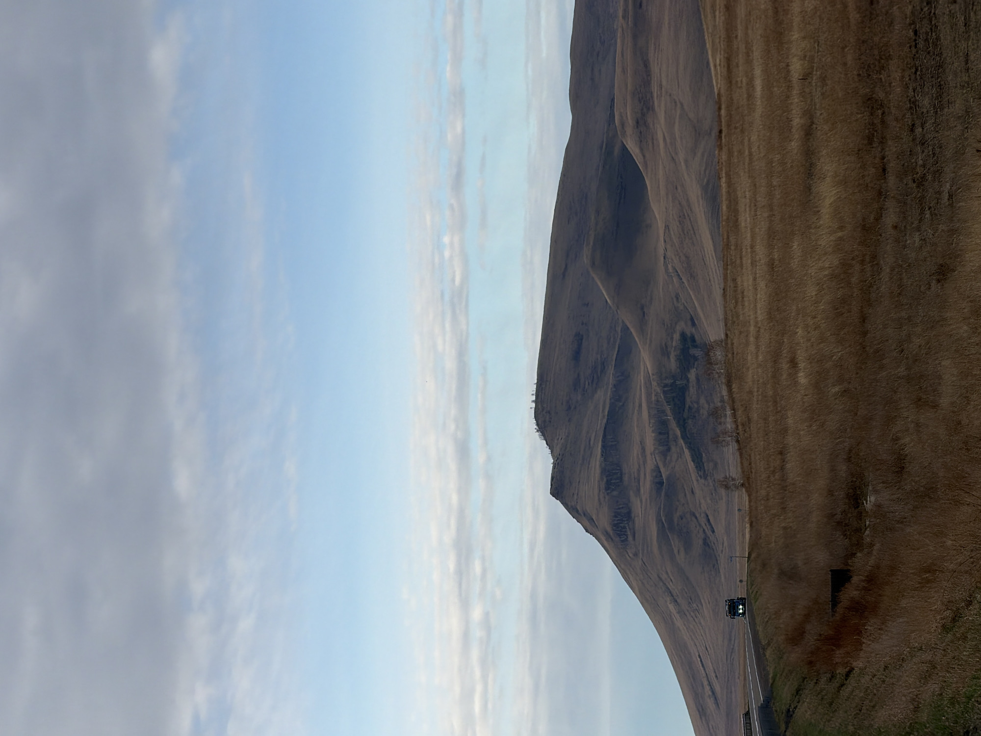 A landscape photograph taken in Сарагаш, Russia, features a steep, brown mountain that dominates the right side of the frame. The mountain's surface is textured with ridges and valleys, suggesting erosion. The lower portion of the mountain transitions into a field of dry, golden-brown grass. A narrow road with a vehicle traveling on it can be seen near the base of the mountain. To the left of the mountain, the sky fills the remainder of the image, showcasing a mix of light blue sky and wispy, white clouds. The photograph is oriented vertically.