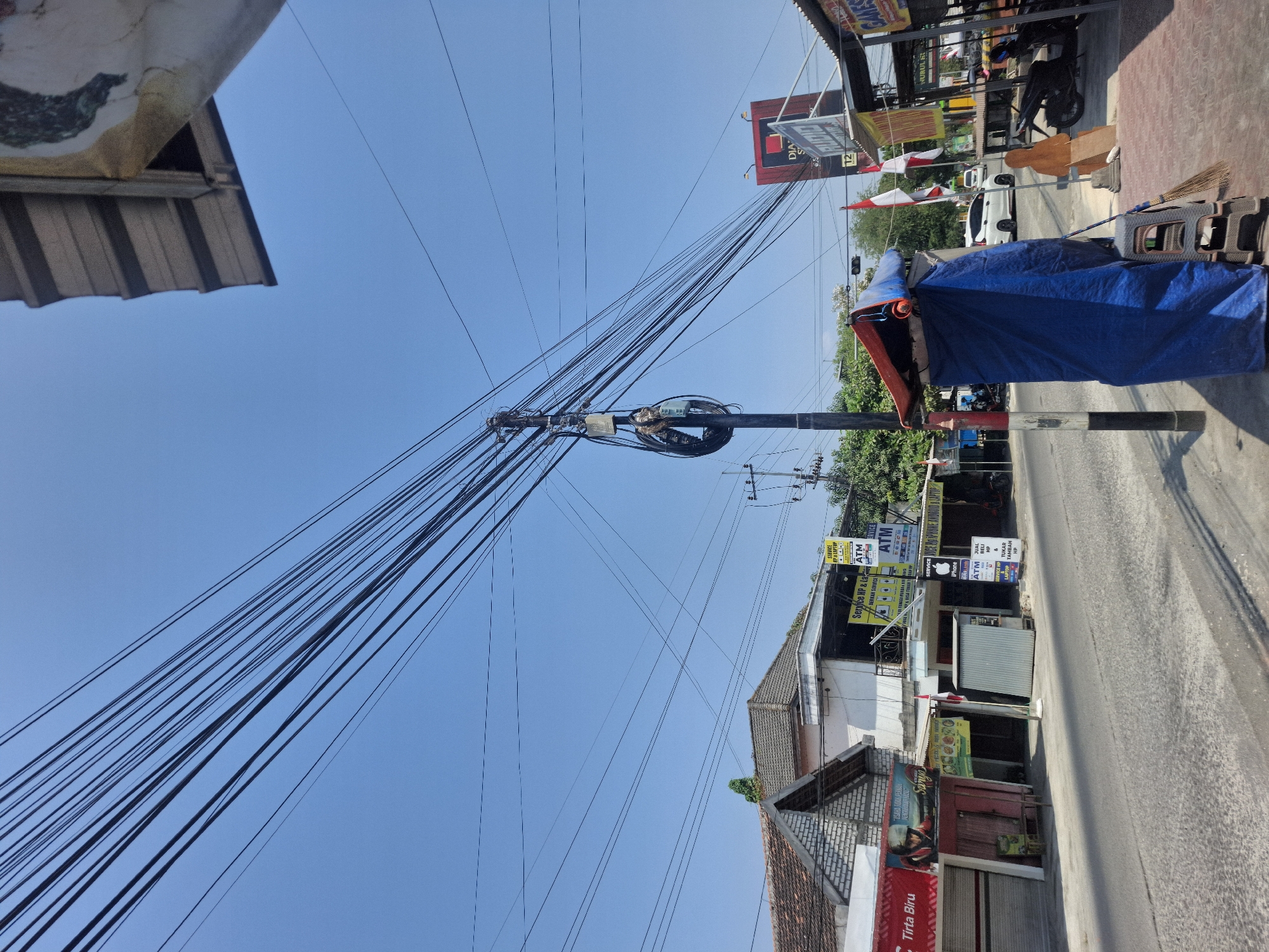 The image shows a street scene in Sumberwudi, Indonesia, under a clear blue sky. A prominent feature is a utility pole overloaded with numerous black electrical and communication wires, some of which are tangled and looped. To the left of the pole, several buildings line the street, including a two-story structure with a tiled roof and white walls, and to its left, a smaller building with a red awning and a closed storefront. Signs are visible on the buildings, including "Service HP & Laptop," "ATM," and "Tirta Biru." In front of one of the buildings, a small silver kiosk stands on the sidewalk.
To the right of the utility pole, a makeshift structure covered with a blue tarp and an orange roof is erected on the sidewalk, with a plastic stool and a broom leaning against it. Further to the right, a tall red billboard with white text is visible. Flags, including the Indonesian flag, are displayed in front of some establishments. A white car is seen driving on the street, and motorcycles are parked on the sidewalk in the background. The road surface appears to be asphalt, and the sidewalks are paved with bricks and concrete. The overall environment suggests a commercial or residential area with active infrastructure and ongoing daily life.
