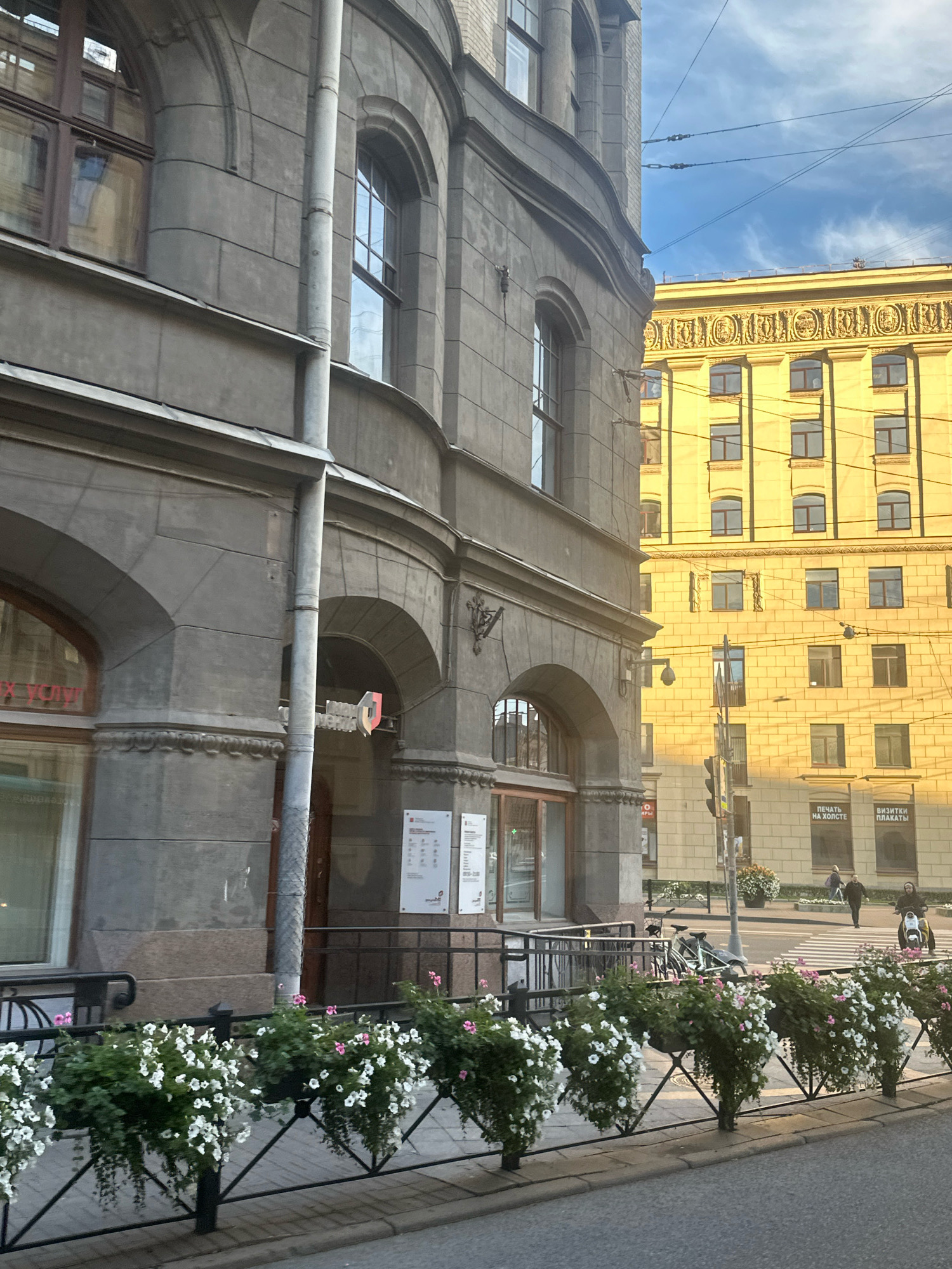 The image depicts a street scene in Saint Petersburg, Russia, on a sunny day with some clouds. In the foreground, a black metal fence is adorned with numerous planters overflowing with white and pink petunias. Behind the fence, a grey stone building with arched windows and decorative elements is visible. A downspout runs vertically along the building's facade. To the right of the grey building, a bright yellow building with numerous windows and ornate architectural detailing stands out. The yellow building is illuminated by the sun, casting a warm glow. Power lines are strung across the sky, crisscrossing above the buildings. In the middle ground, a crosswalk is visible on the street, with two individuals walking across it. Further to the right, a person is riding a scooter. Bicycles are parked on the sidewalk near the grey building. Several signs are posted on the buildings, with some text visible in Russian. The overall impression is an urban environment with classic architecture and vibrant floral decorations.