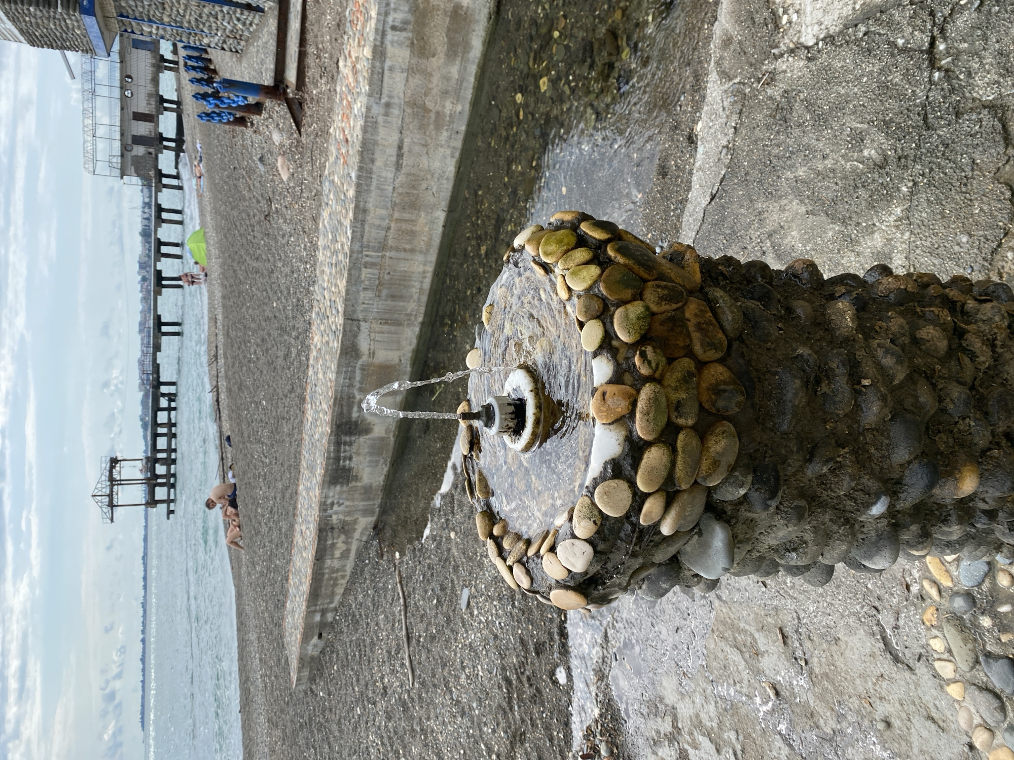 The image depicts a seaside scene in Sokhumi, Abkhazia, featuring a small decorative fountain in the foreground. The fountain is made of concrete and adorned with rounded, multi-colored pebbles. Water streams from its center, creating a gentle spray. In the background, a pebble beach slopes towards the Black Sea. A pier with a covered structure extends into the water, and a building with a white upper level and a stone-clad lower level is visible on the right. Several people are present on the beach; two individuals are lying down near the water's edge, while others are further back, some near a green tent. The sky is overcast with scattered clouds.