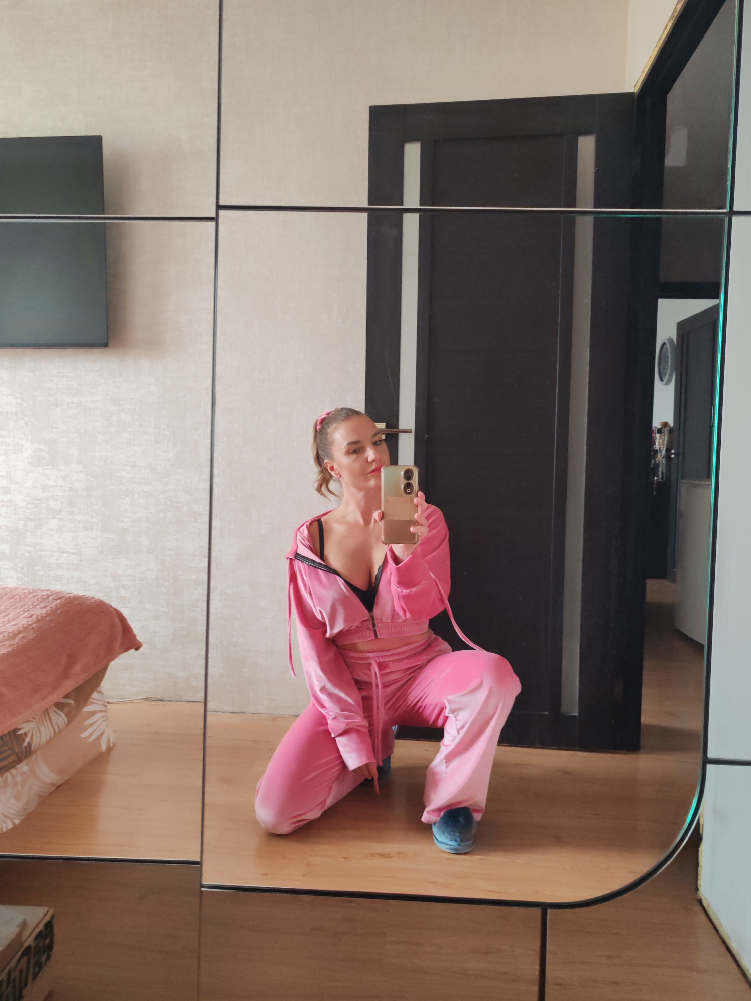 A fair-skinned woman with her hair in a ponytail, wearing a pink velour tracksuit and dark bra, kneels and takes a selfie in front of a large mirror. The mirror reflects a sparsely furnished room with a bed and a dark door leading to another room. The room’s location is unknown and cannot be determined to be Petropavlovsk-Kamchatsky, Russia, based on the visual data.