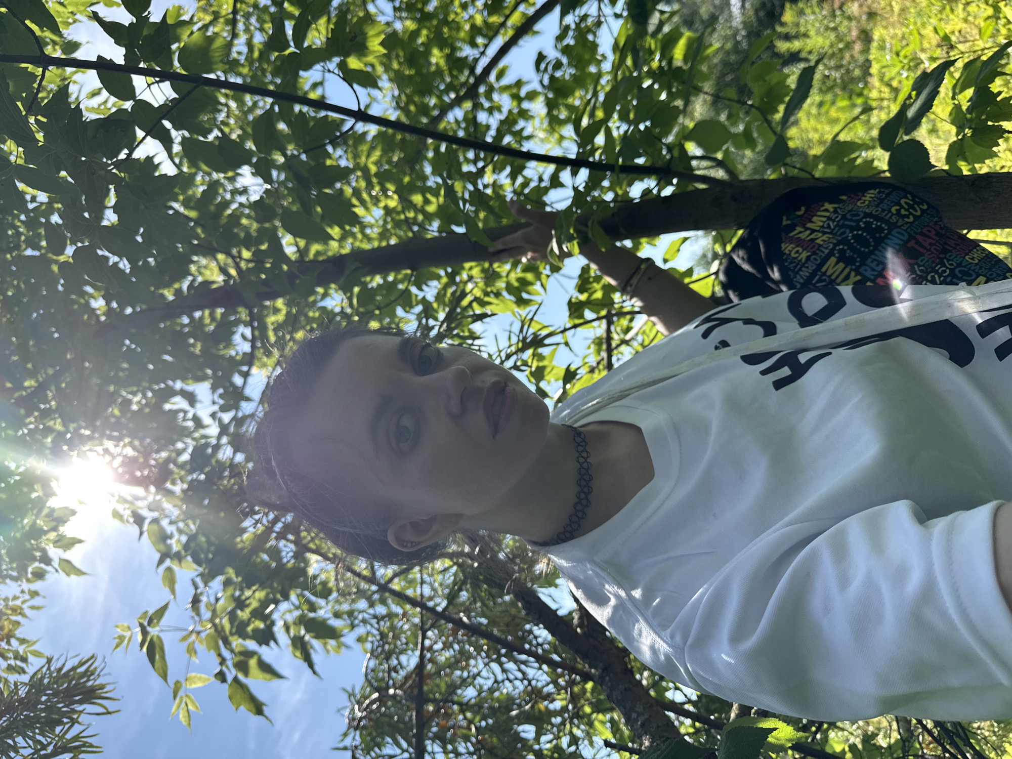 A low-angle medium shot captures a young person with dark hair pulled back in a bun, looking towards the camera while standing amongst trees. The person wears a white t-shirt with a black graphic and a black choker necklace. Their right arm is extended, touching a tree trunk, while their left arm is partially visible. Sunlight filters through the green leaves, creating lens flare and highlighting the bright blue sky. The background consists of more trees and foliage, suggesting an outdoor, possibly wooded, environment. The location is unknown, within a city or area identified as Unknown.