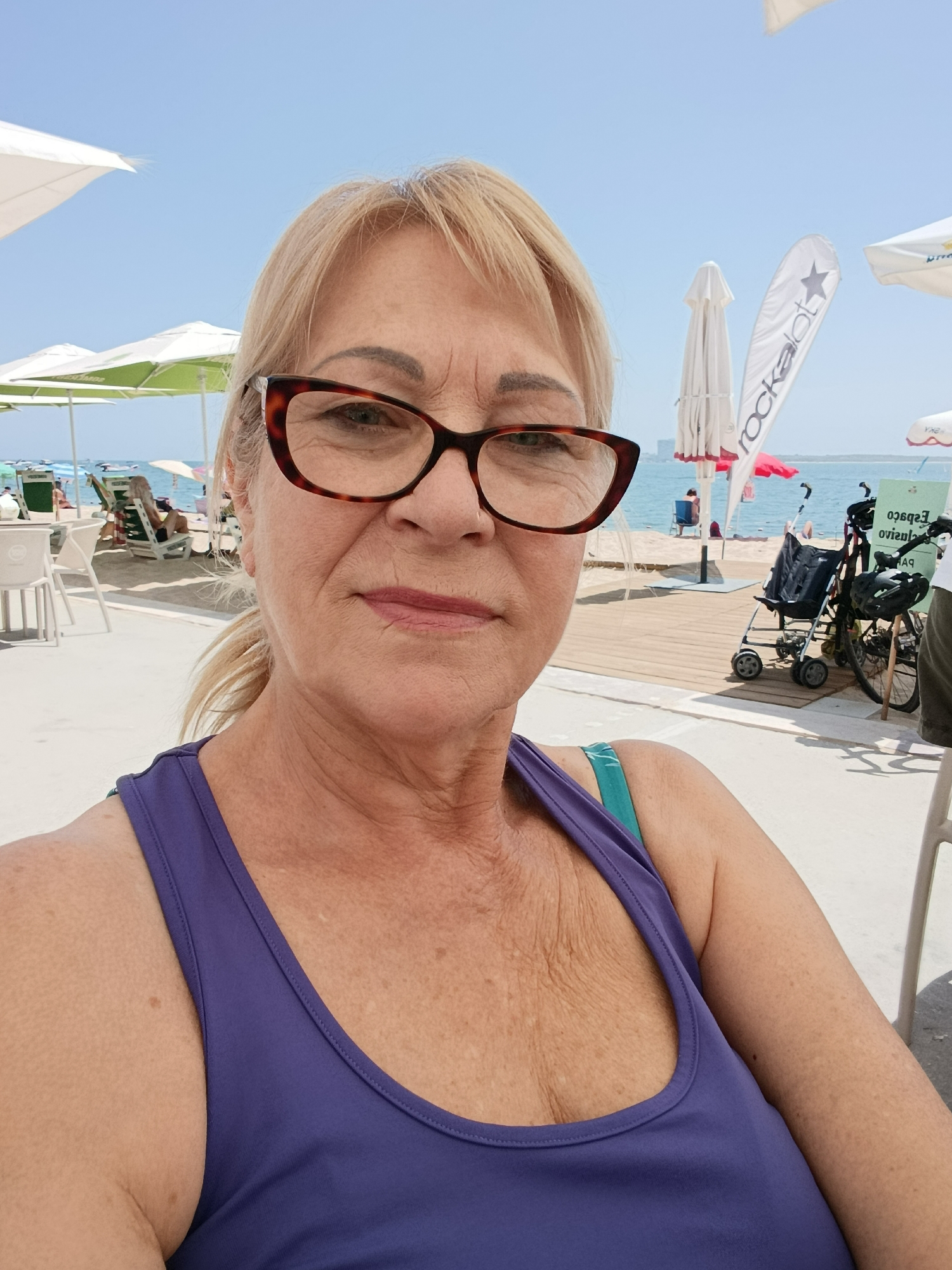 The image shows a woman wearing glasses and a purple tank top, taking a selfie on a sunny day at a beach. She has blonde hair pulled back and is looking at the camera. In the background, there are beach umbrellas, lounge chairs, people relaxing on the sand, and the ocean. There is also a wooden boardwalk with a stroller and a bicycle. The scene appears to be a beachside cafe or restaurant in Setúbal, Portugal, possibly near Praia da Figueirinha or Praia do Portinho da Arrábida.