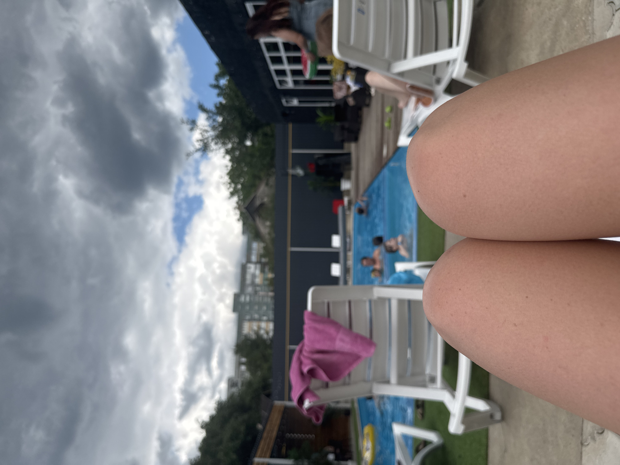 An overhead view from a reclining perspective shows a portion of two human legs in the foreground. The legs are smooth and tanned, with a scattering of small freckles visible on the skin. Behind the legs, a swimming pool area is visible. Several white lounge chairs are positioned around the pool. One chair in the mid-ground has a pink towel draped over its back. In the pool, several people are swimming and playing. A yellow inflatable ring is floating in the pool. To the right, a dark building with white-framed windows is present, and a woman in a light blue dress is seen carrying a round, red object, possibly a frisbee or a serving tray with watermelon slices. In the background, trees and multi-story buildings are visible under a cloudy sky. The overall scene suggests a leisure setting, possibly a hotel or a resort, with a focus on a swimming pool.