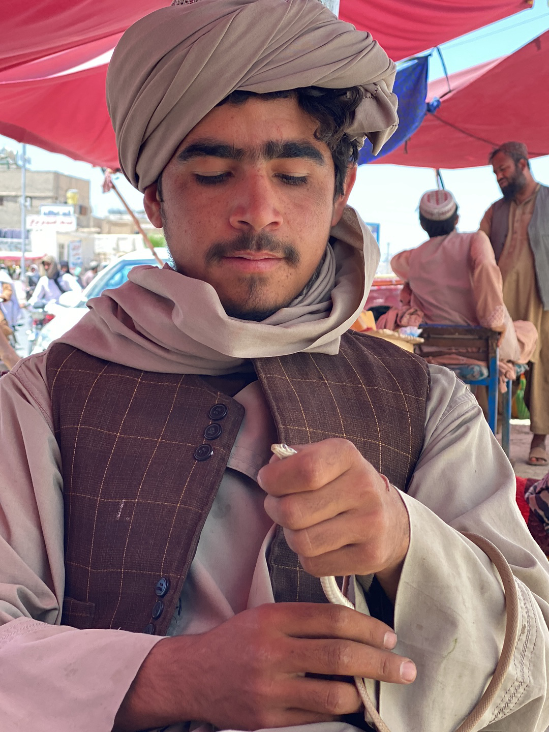 A man wearing a turban and vest holds a small, light-colored snake in his hands. He is situated outdoors under a red and blue canopy. In the background, there are other men, some seated, along with pedestrians and vehicles, suggesting a city street scene. The image was taken in the city of قندهار, Afghanistan.