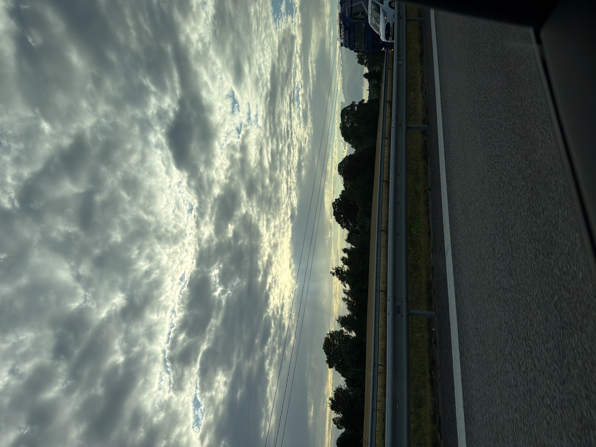 The image captures a view from a vehicle traveling on a highway, likely near Himle, Sweden, given the context provided. The sky is predominantly filled with heavy, gray clouds, with breaks allowing shafts of sunlight to pierce through, illuminating portions of the horizon and creating a dramatic, atmospheric effect. Below the sky, a line of trees forms a dark silhouette against the lighter sky. A metal guardrail separates the highway from the grassy embankment and fields beyond. In the distance, on the highway, a blue truck with a white cab is visible, along with a white vehicle in front of it. The road surface is asphalt with a white lane marker. The perspective suggests the photo was taken while in motion.